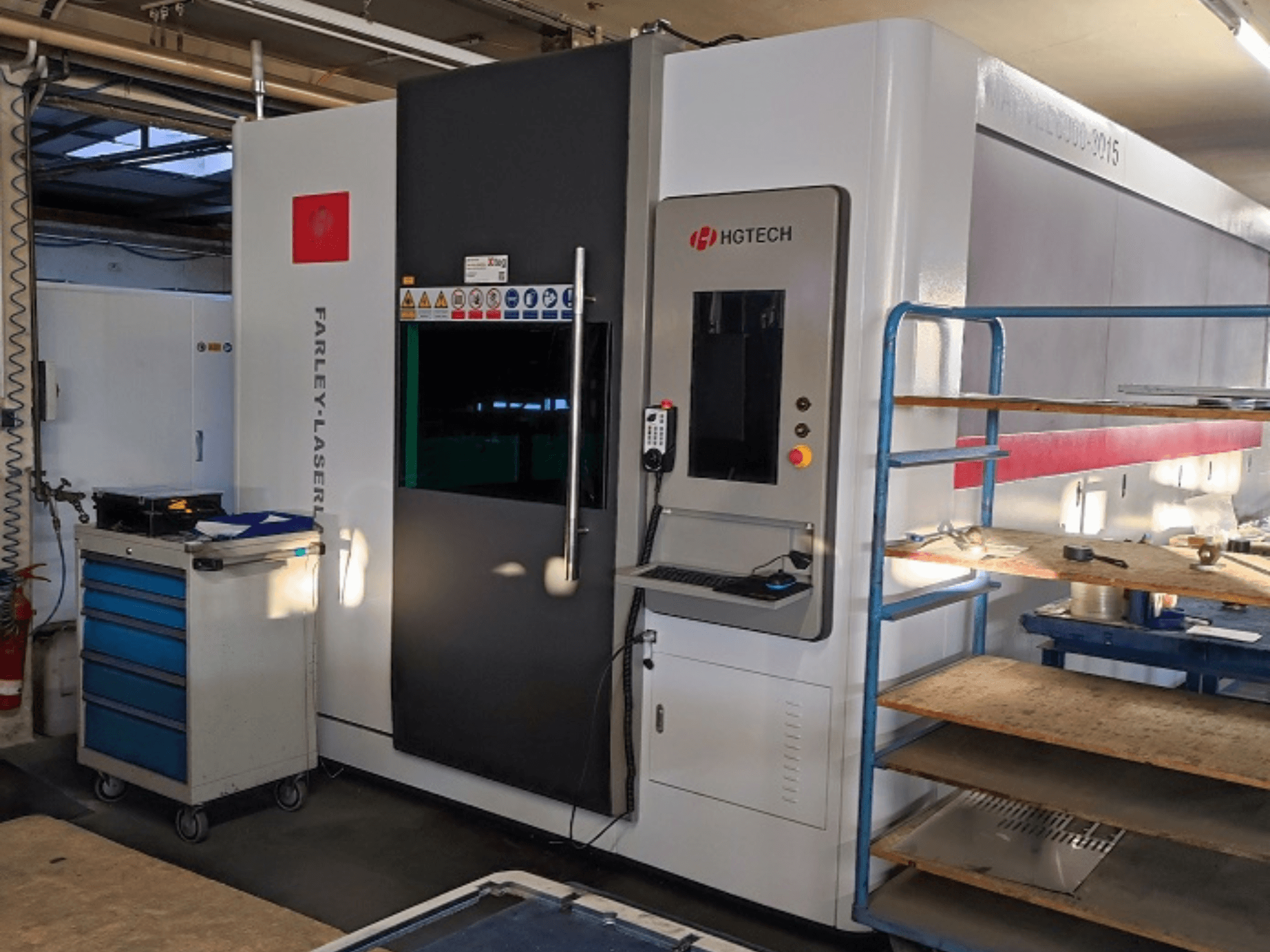 Máquina de corte a laser de fibra HGTECH Marvel 6000-3015, vista frontal com painel de controlo e armazenamento adjacente.