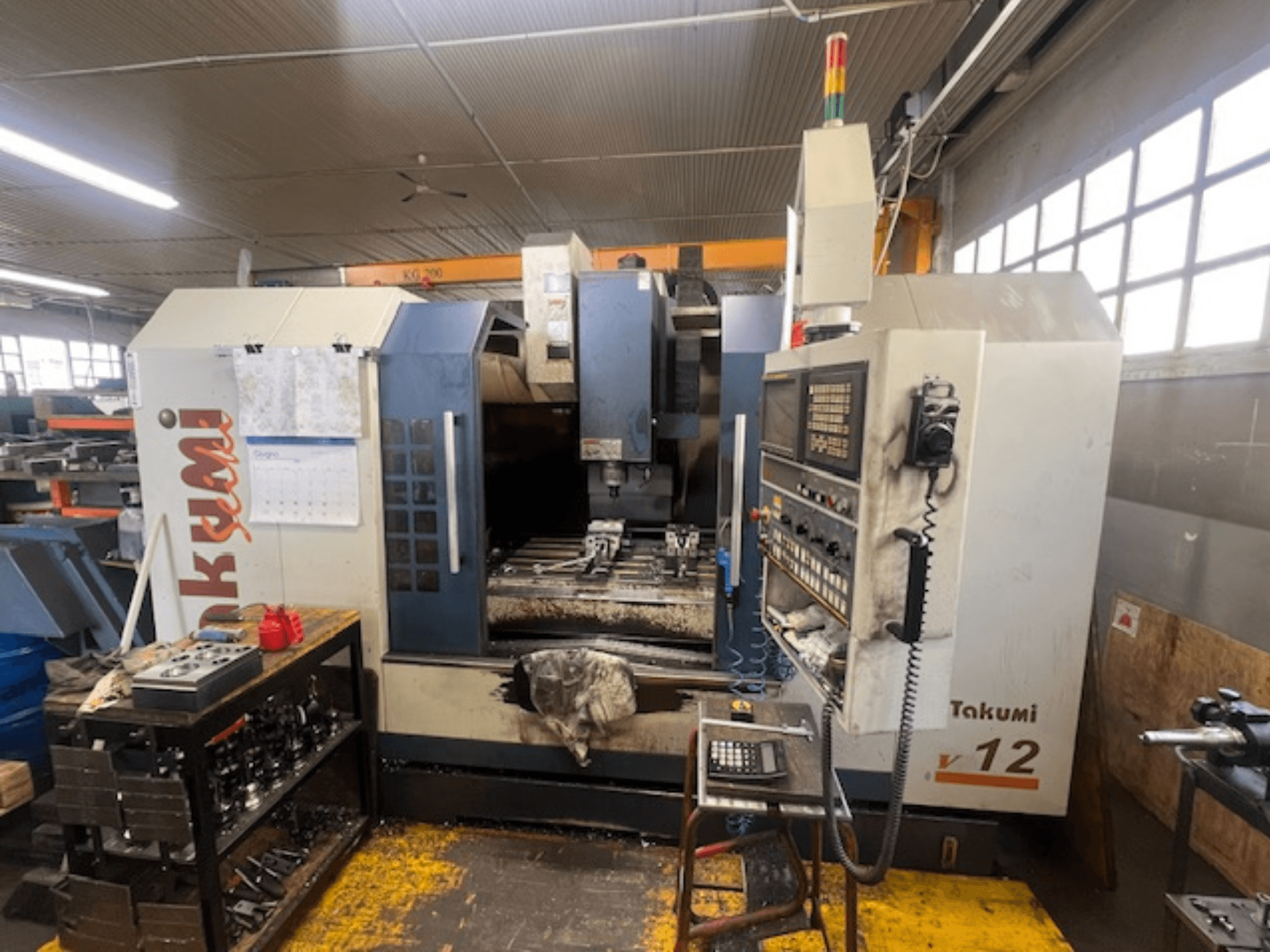 Fresadora CNC Takumi V12 em vista lateral, mostrando o painel de controlo, a área de corte e as ferramentas na estação de trabalho.