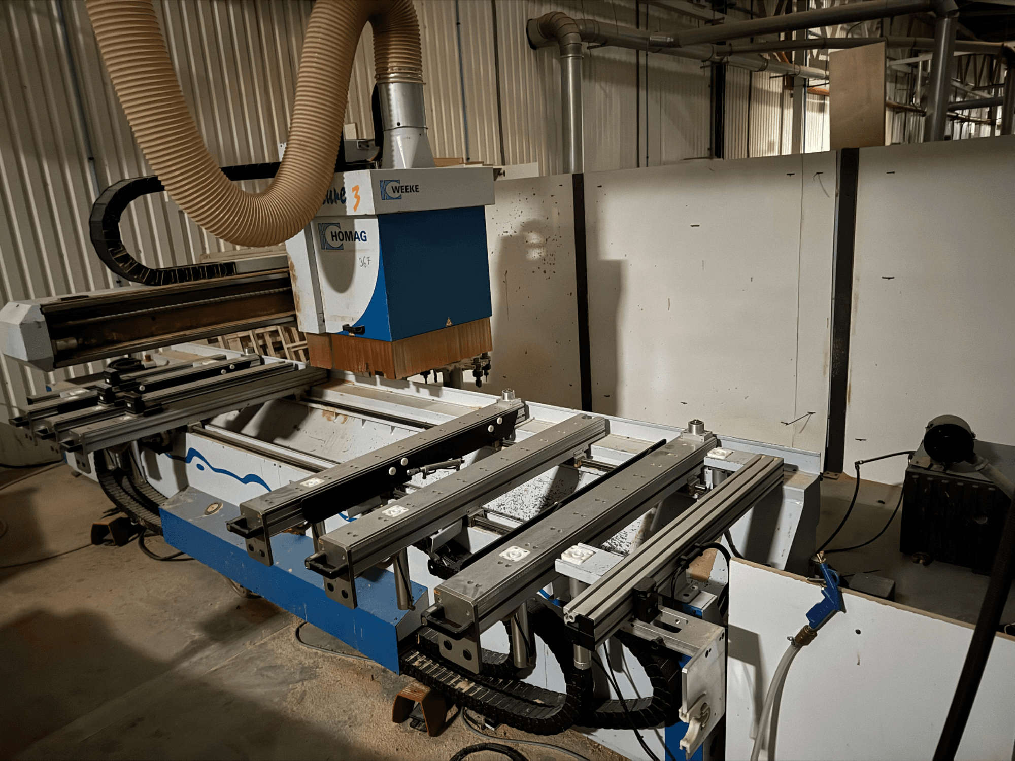 Máquina CNC HOMAG de design azul e branco, vista lateral, com calhas deslizantes, sistema de extração de poeiras e ferramentas montadas.