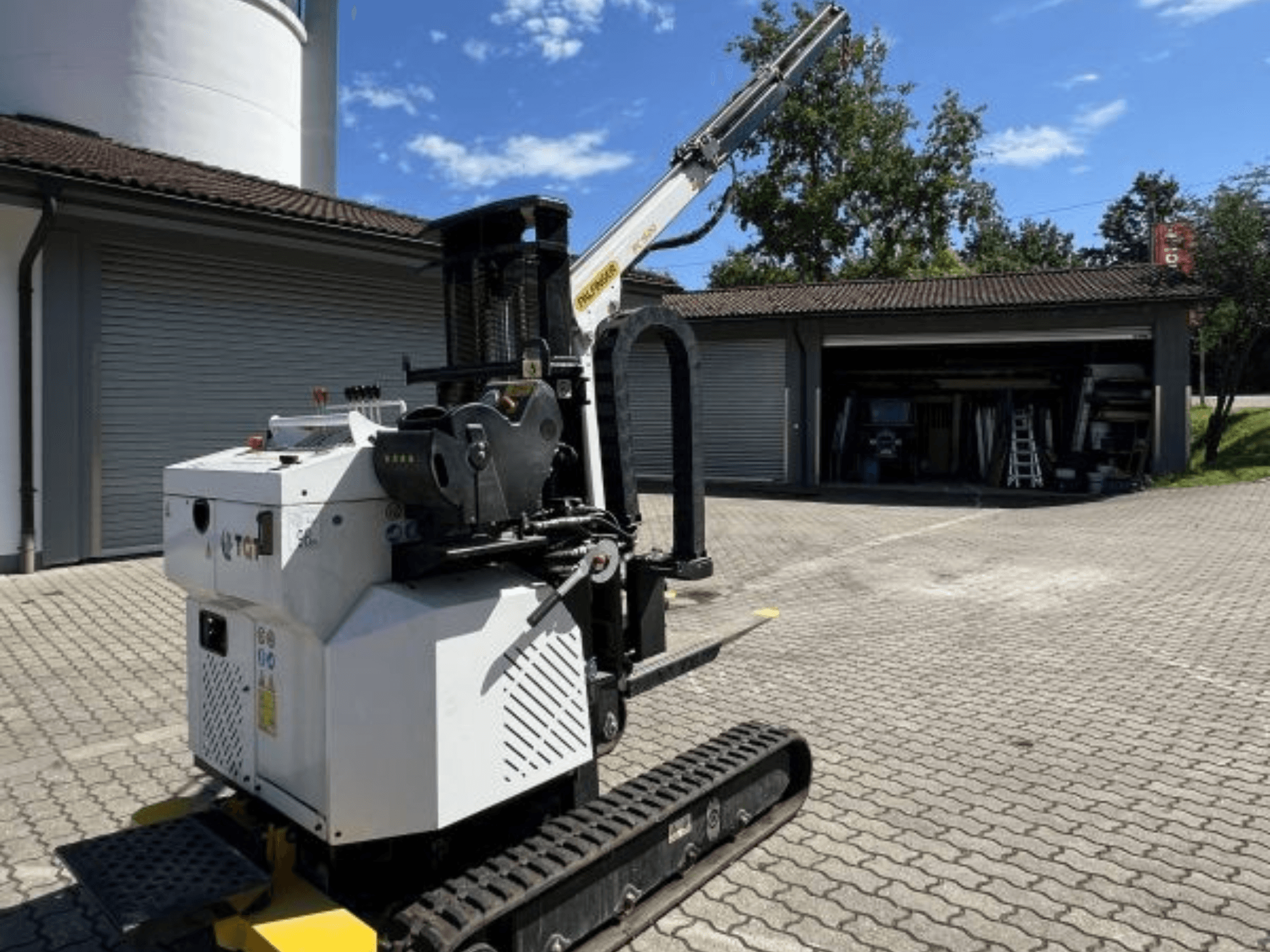 Mini grua de construção TGI, vista lateral, mostrando os carris e a lança montada, estacionada numa superfície de tijolo junto a uma garagem.