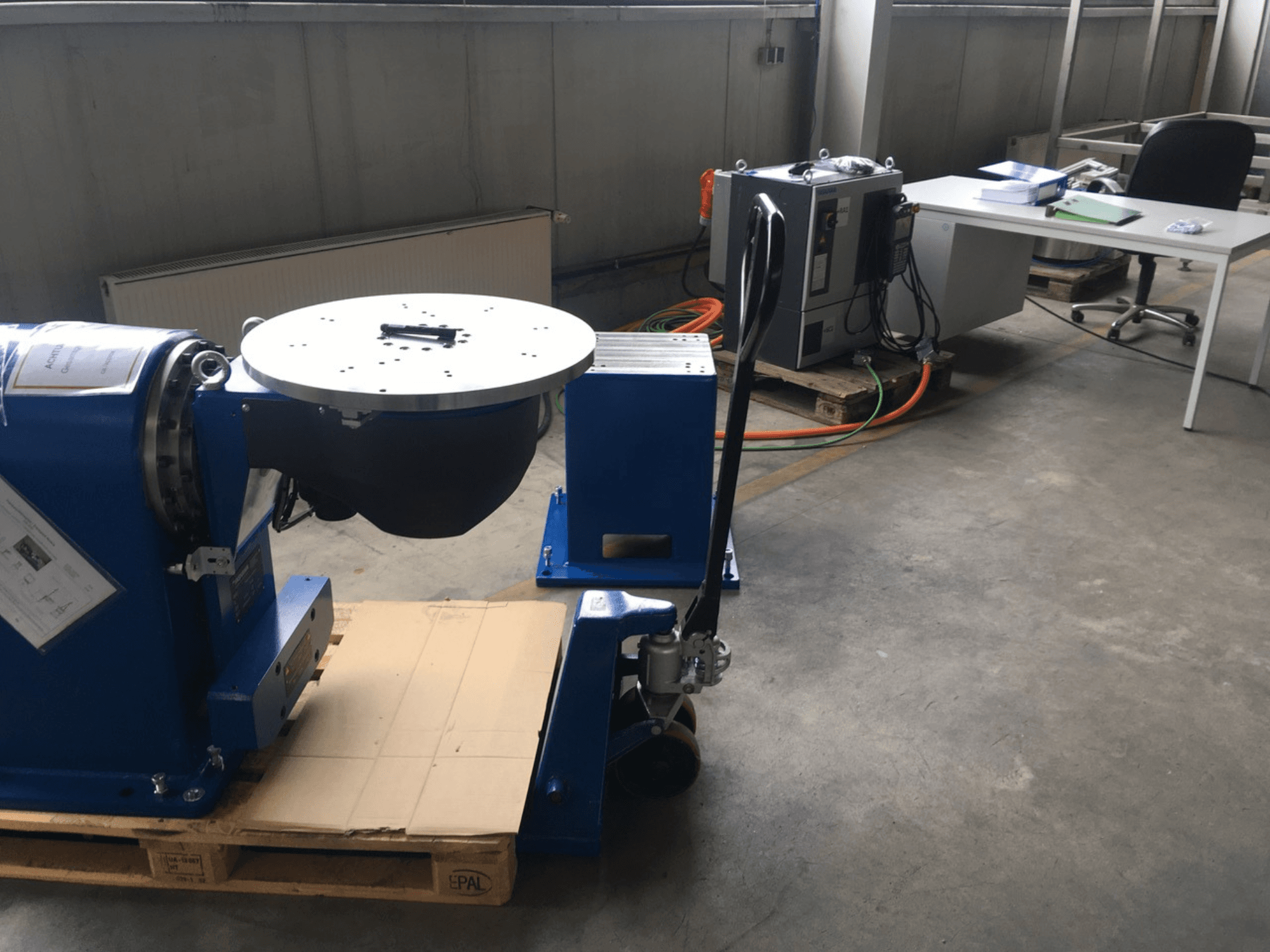 Equipamento industrial azul com uma base circular e um painel de controlo, visto de frente, numa palete de madeira num ambiente de escritório.