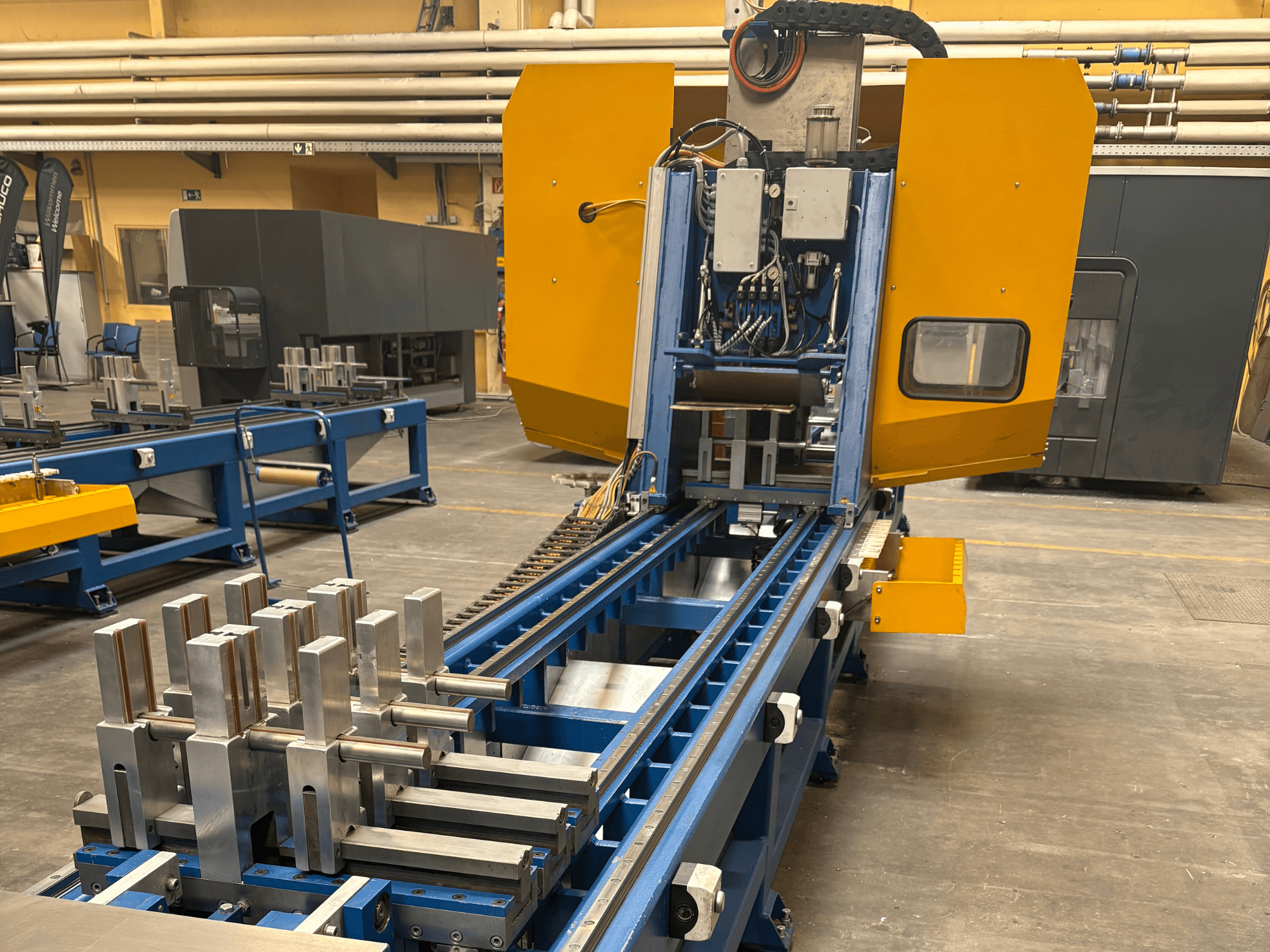 Vista frontal do Centro de Maquinação Universal SCHÜCO PBX AL, mostrando peças metálicas numa mesa de maquinação azul.