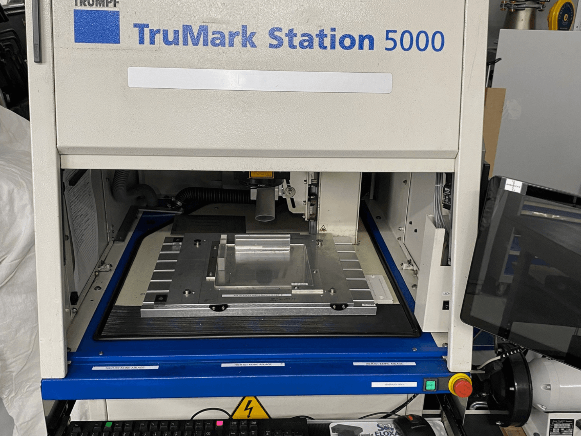 Vista frontal da máquina de marcação a laser TruMark Station 5000, mostrando a mesa de trabalho, os comandos e o ecrã de monitorização.