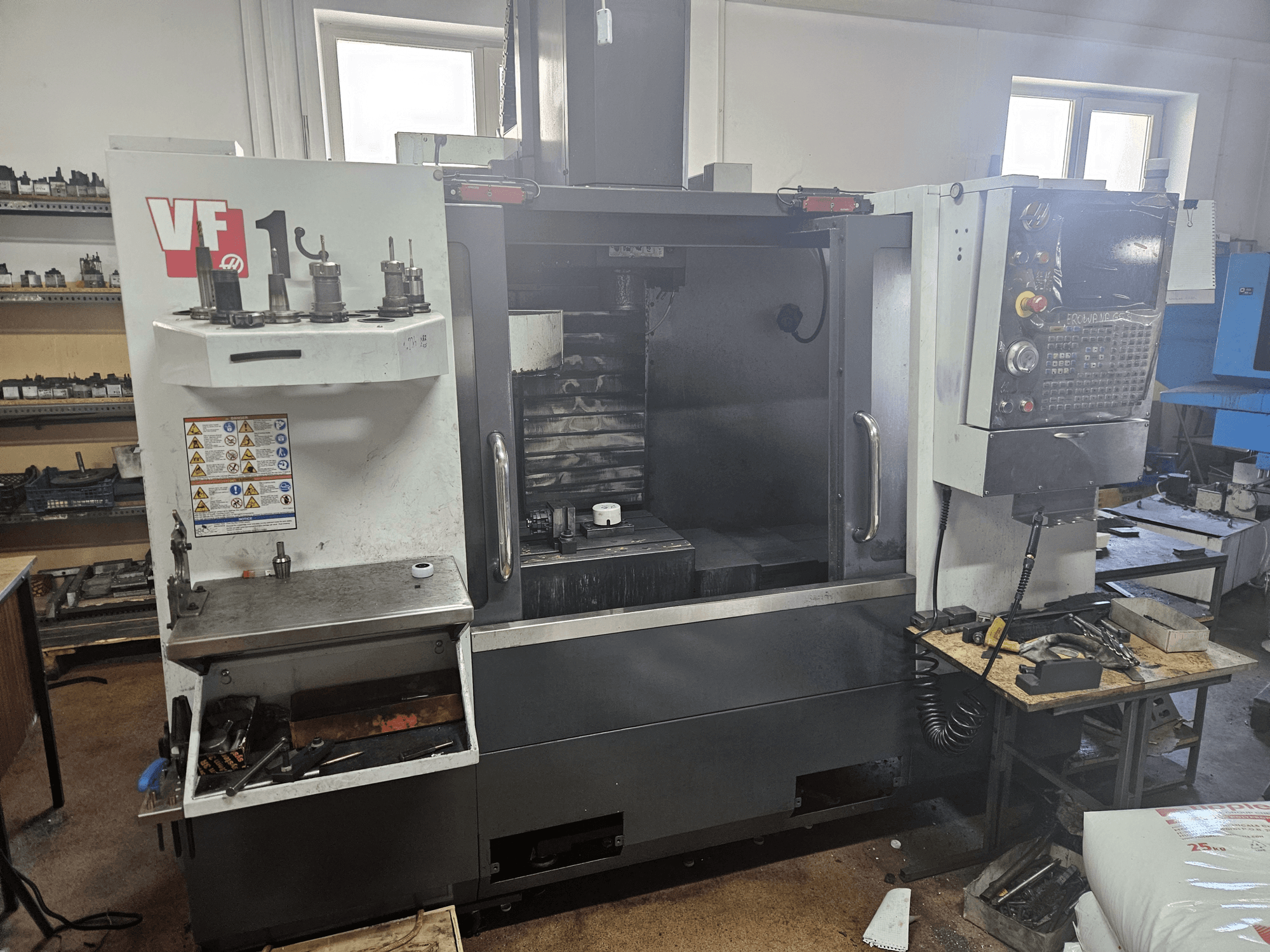 Centro de maquinação vertical HAAS VF-1, vista frontal com ferramentas de lado, painel de controlo visível e espaço de trabalho em segundo plano.