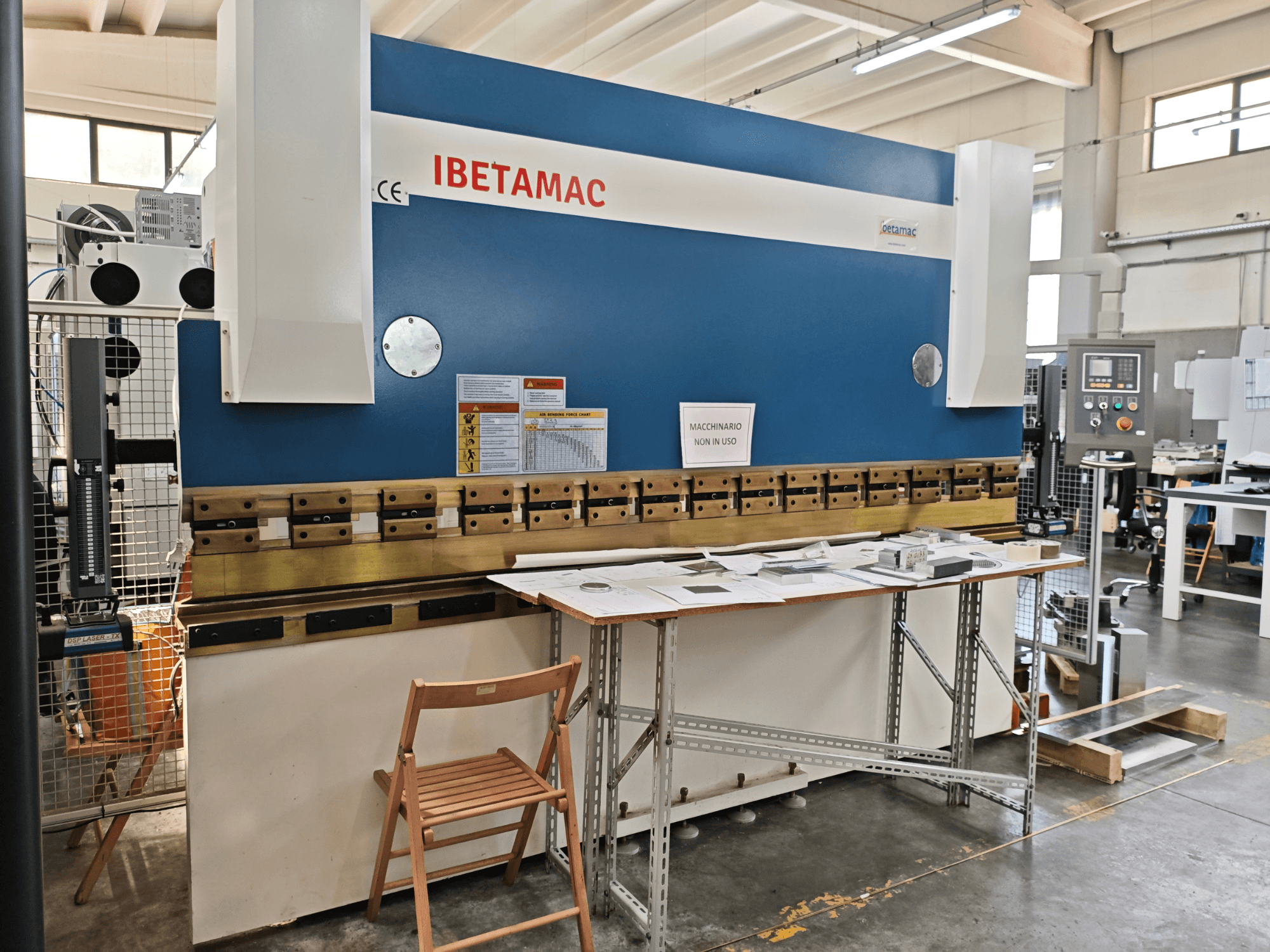 Prensa-dobra IBETAMAC 3200 100T numa oficina, vista lateral que mostra o painel de controlo, a área de trabalho e as indicações de segurança.
