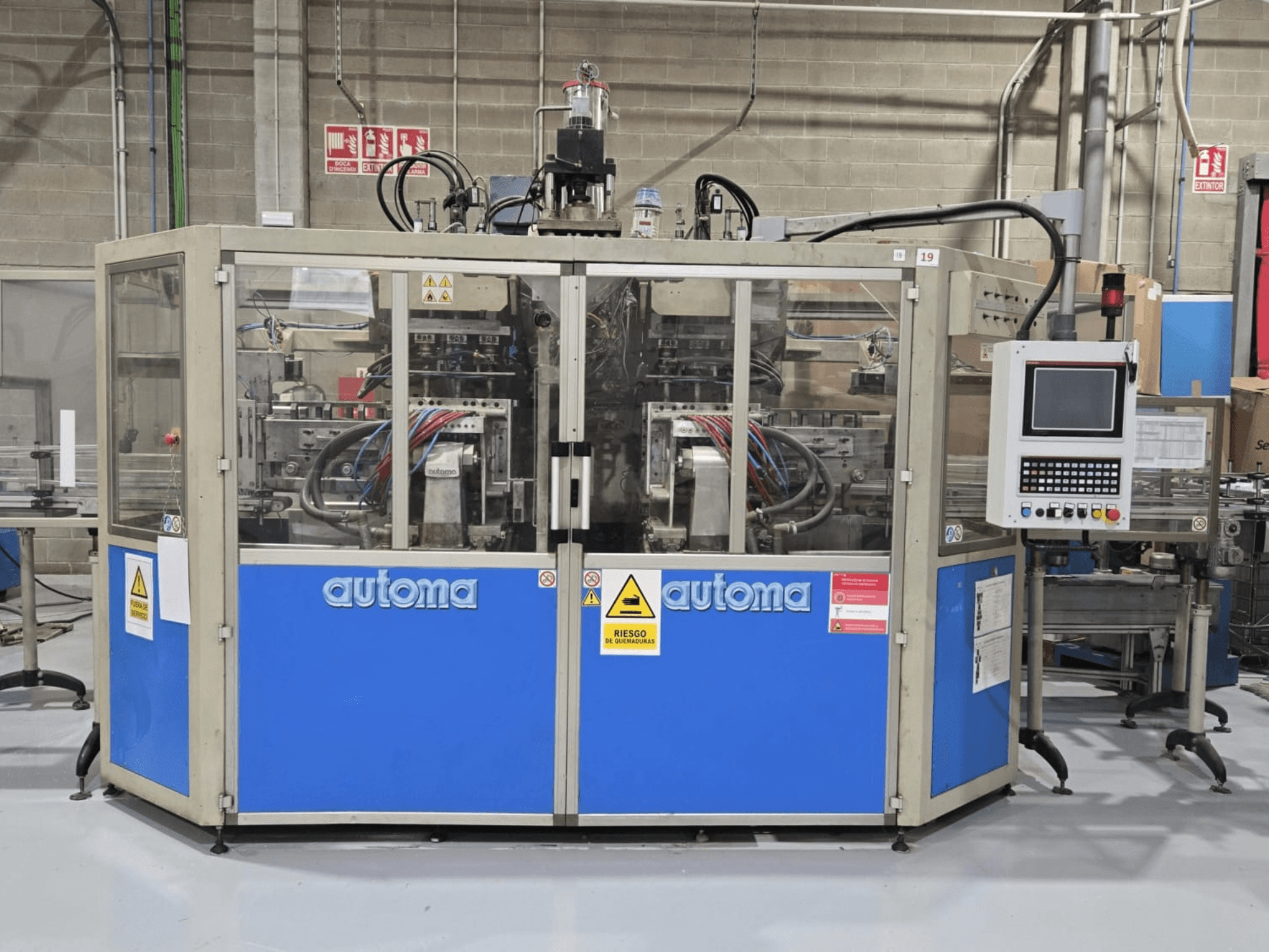 Vista frontal da máquina de moldagem por sopro AUTOMA APEX AT500D, com o painel de controlo e várias mangueiras, instalada num ambiente fabril.