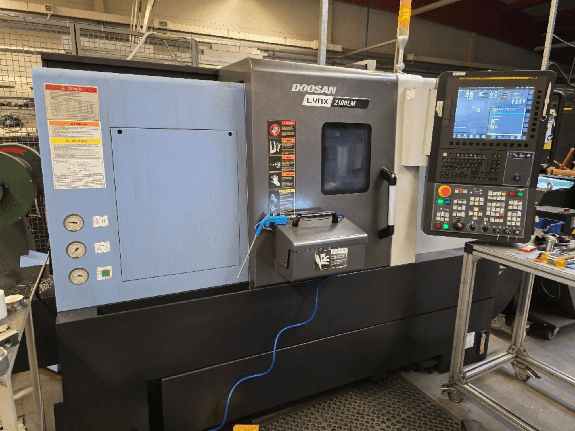 Torno CNC Doosan Lynx 2100LM, vista frontal, com um painel de controlo, manómetros e avisos de segurança.