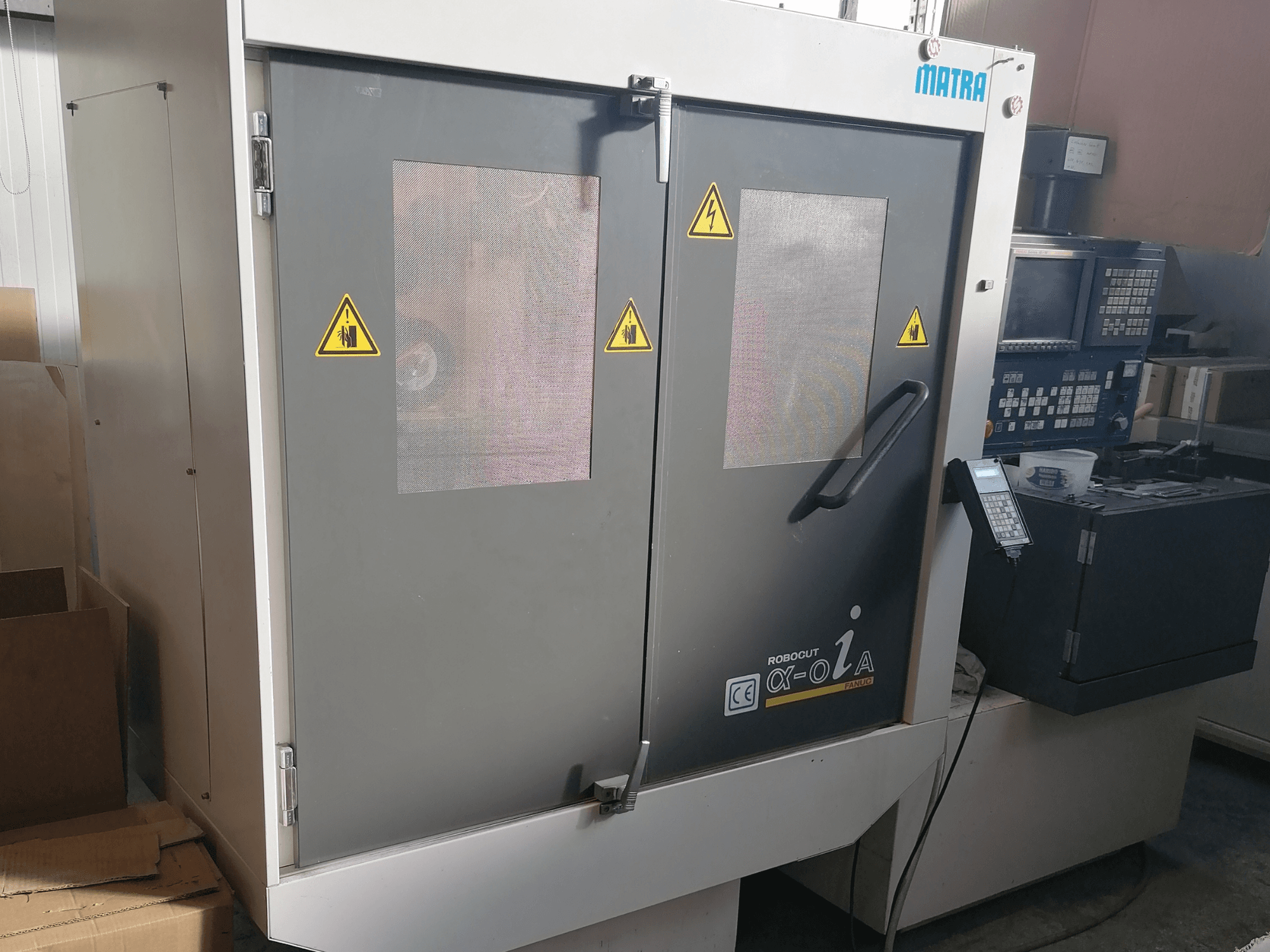 Máquina Robocut α-oZα da Matra, vista frontal com avisos de segurança, painel de controlo e portas duplas com janelas de rede.