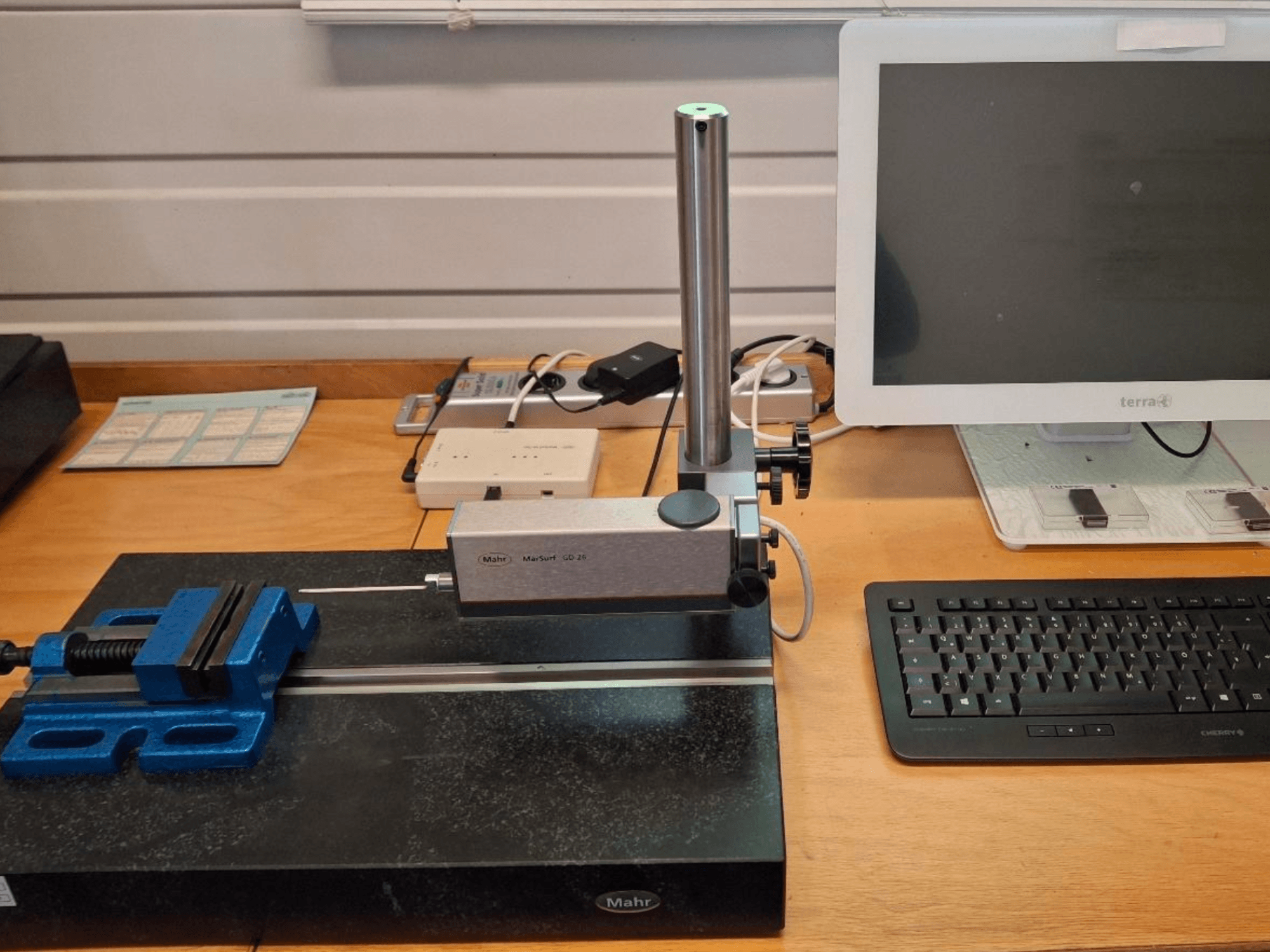MAHR MARSURF GD 26 CMM numa bancada de trabalho, com uma sonda de medição, um torno azul e um monitor de computador. Vista frontal.
