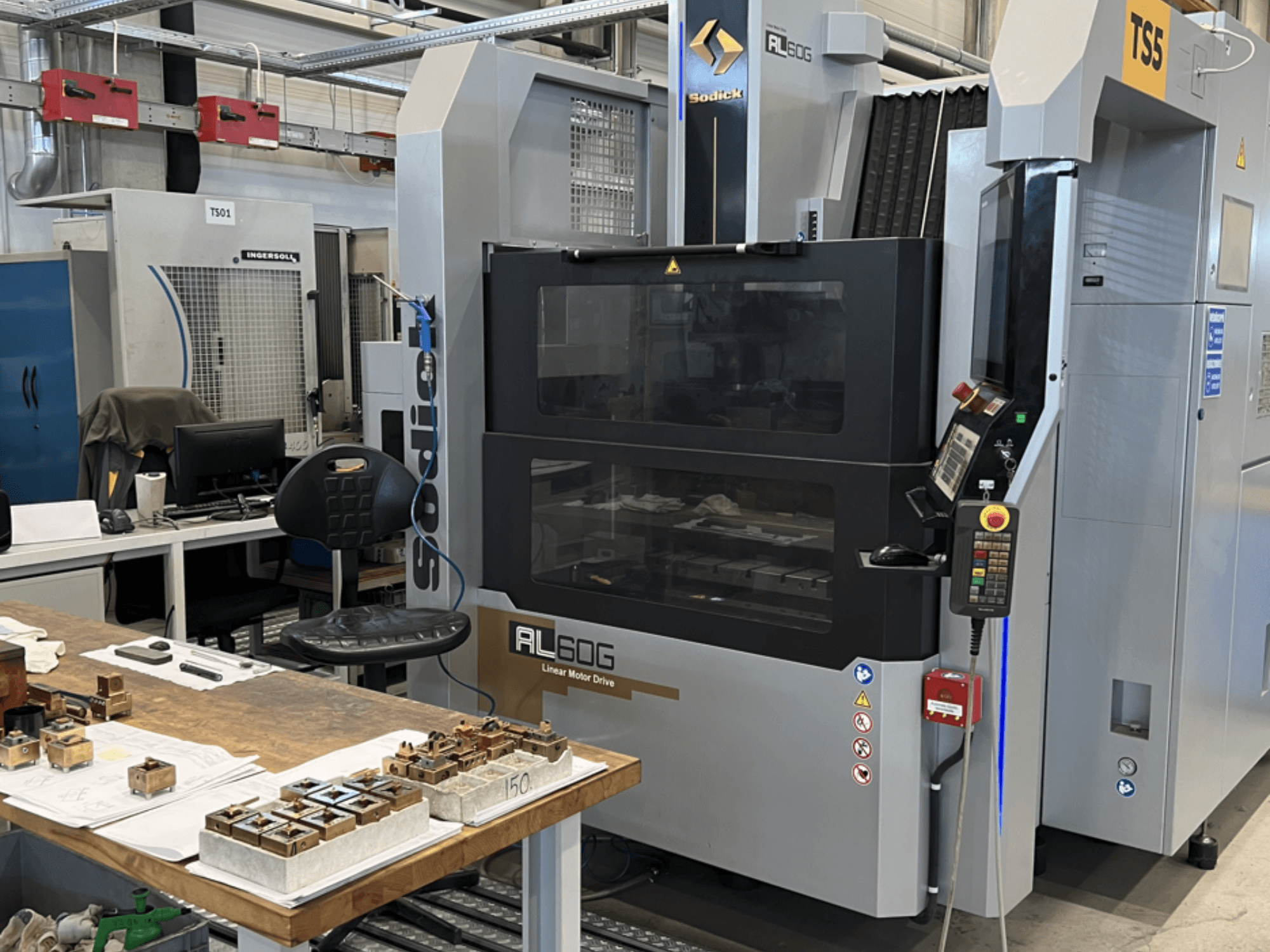 Máquina EDM Sodick AL60G para abrir moldes; vista frontal mostrando os controlos, a janela e o espaço de trabalho com várias peças metálicas.