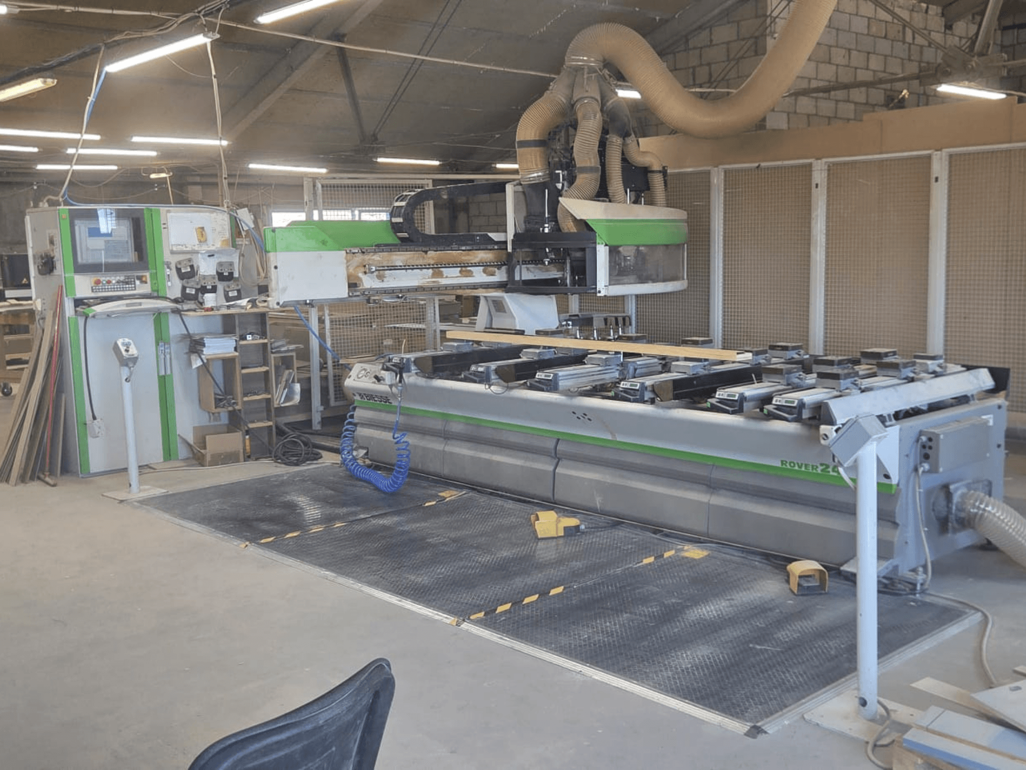 Centro de processamento CNC BIESSE Rover 24S numa oficina, com um painel de controlo e uma grande superfície de trabalho com peças de madeira.