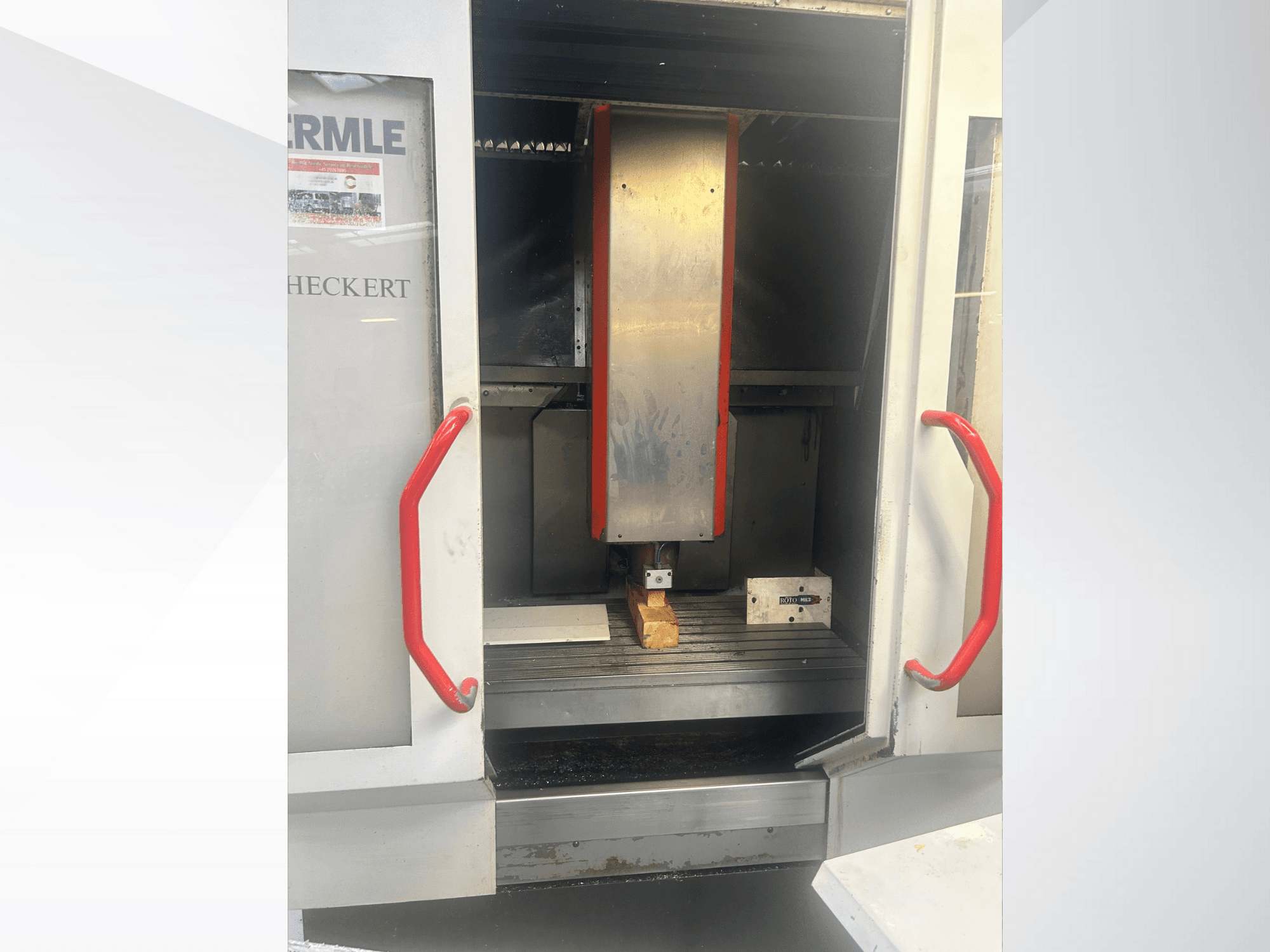 Vista frontal da máquina CNC Hermle, com um interior metálico, pegas vermelhas e uma peça de trabalho sobre a mesa.