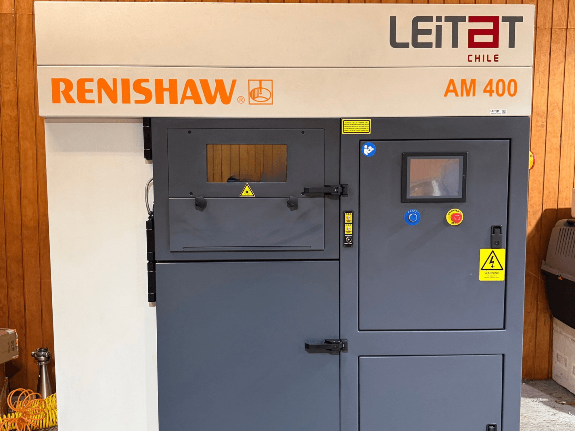 Vista frontal da impressora 3D de metal RENISHAW AM400, com o painel de controlo, as etiquetas de aviso e o acesso para manutenção.