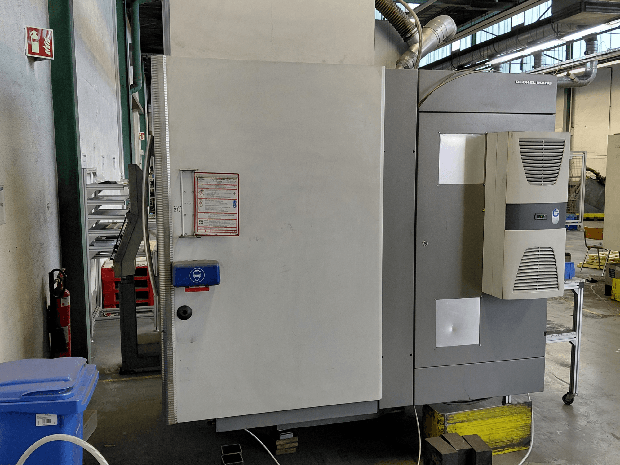 Vista lateral do centro de maquinação vertical DECKEL MAHO DMC 635 V com o painel de controlo e as etiquetas de segurança visíveis.