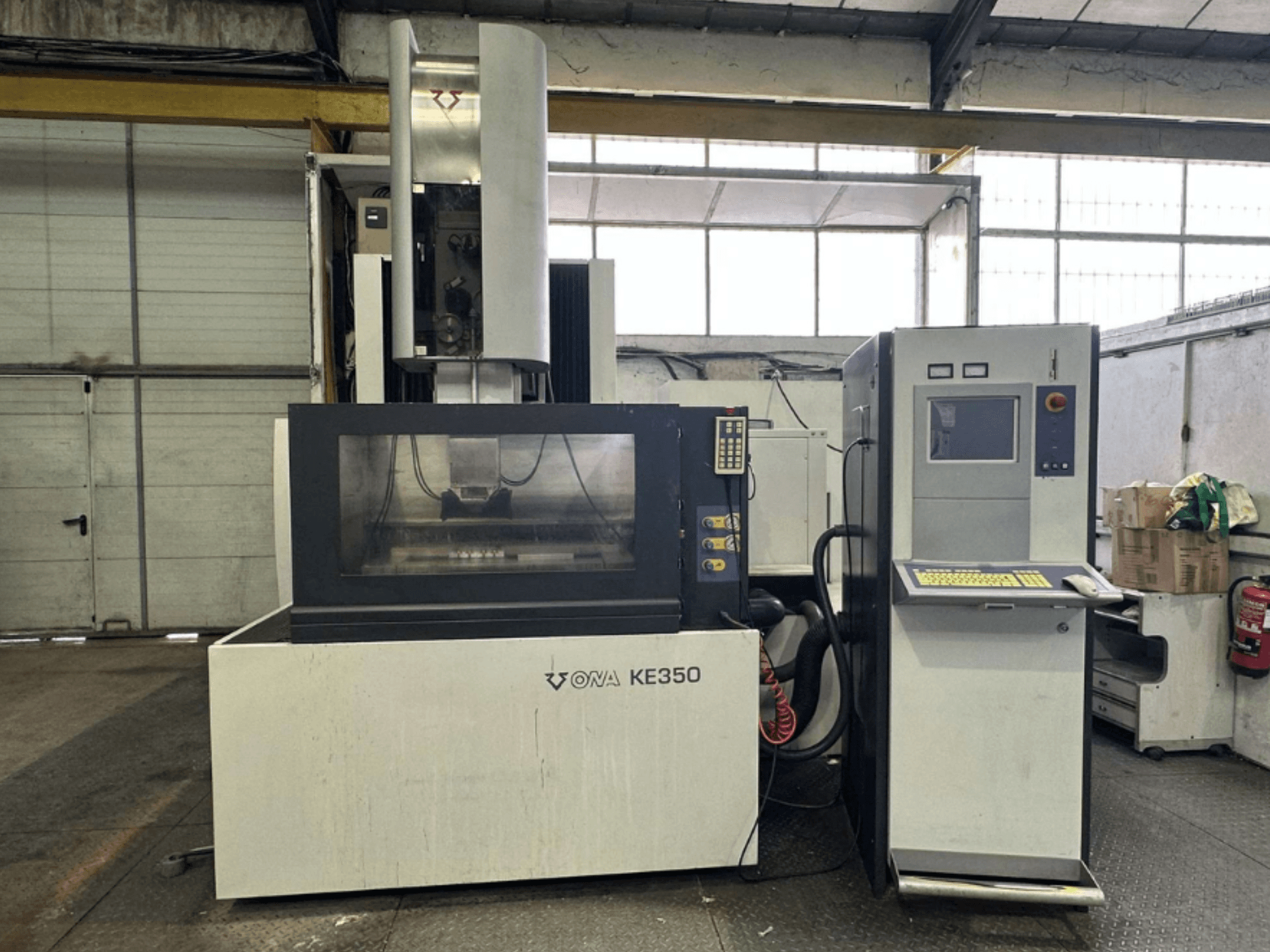 Vista frontal da máquina CNC ONA KE350 com painel de controlo, ecrã de visualização e vários botões, localizada num ambiente industrial.