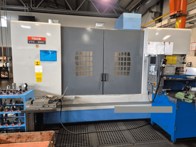 Máquina Mazak VTC-300C numa fábrica, vista frontal mostrando o painel de controlo, portas cinzentas com grelha de janelas e equipamento próximo.