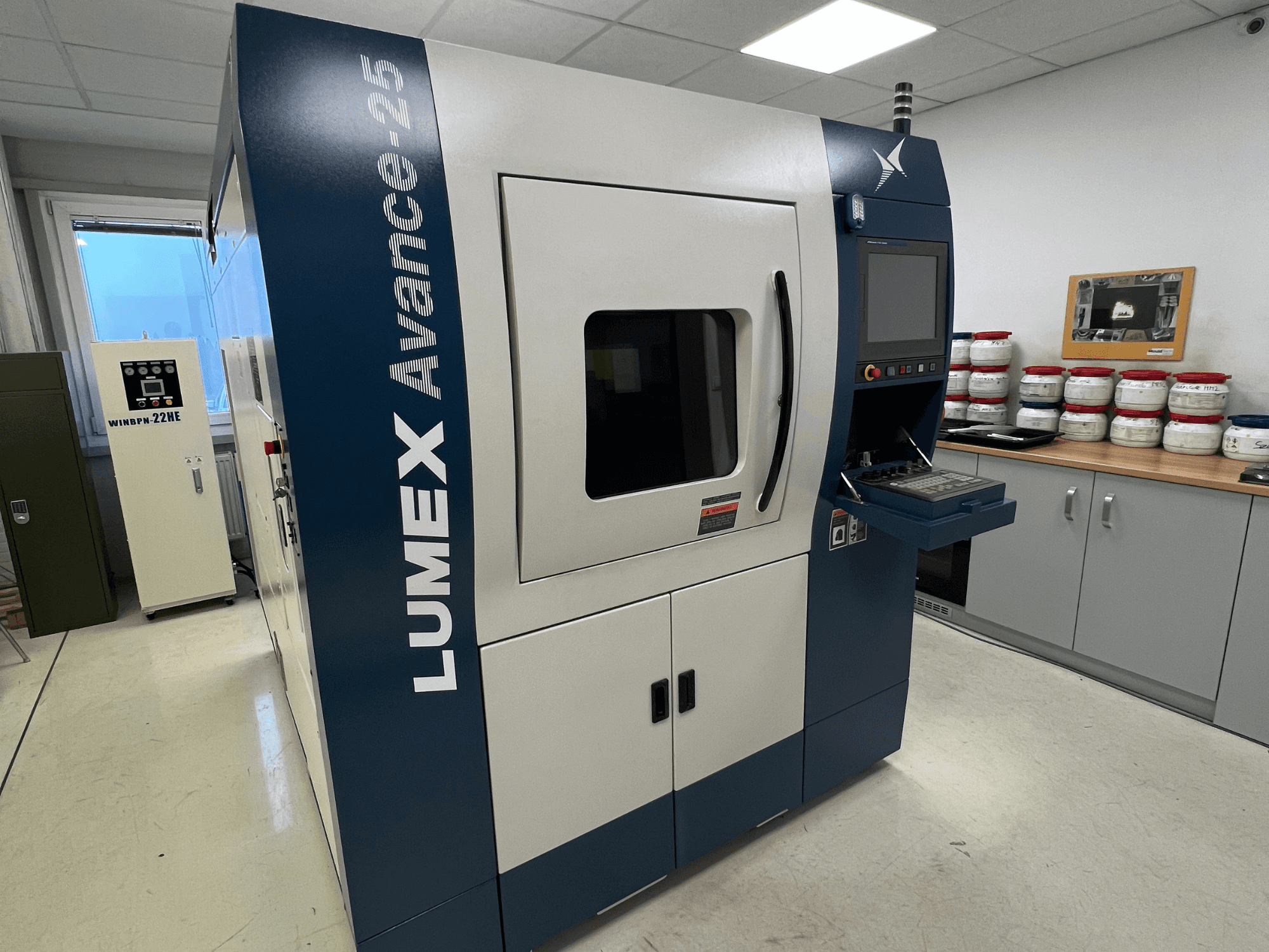 Impressora LUMEX Avance-25 3D num laboratório, vista frontal. Painel de controlo com ecrã e botões visíveis, equipamento em segundo plano.