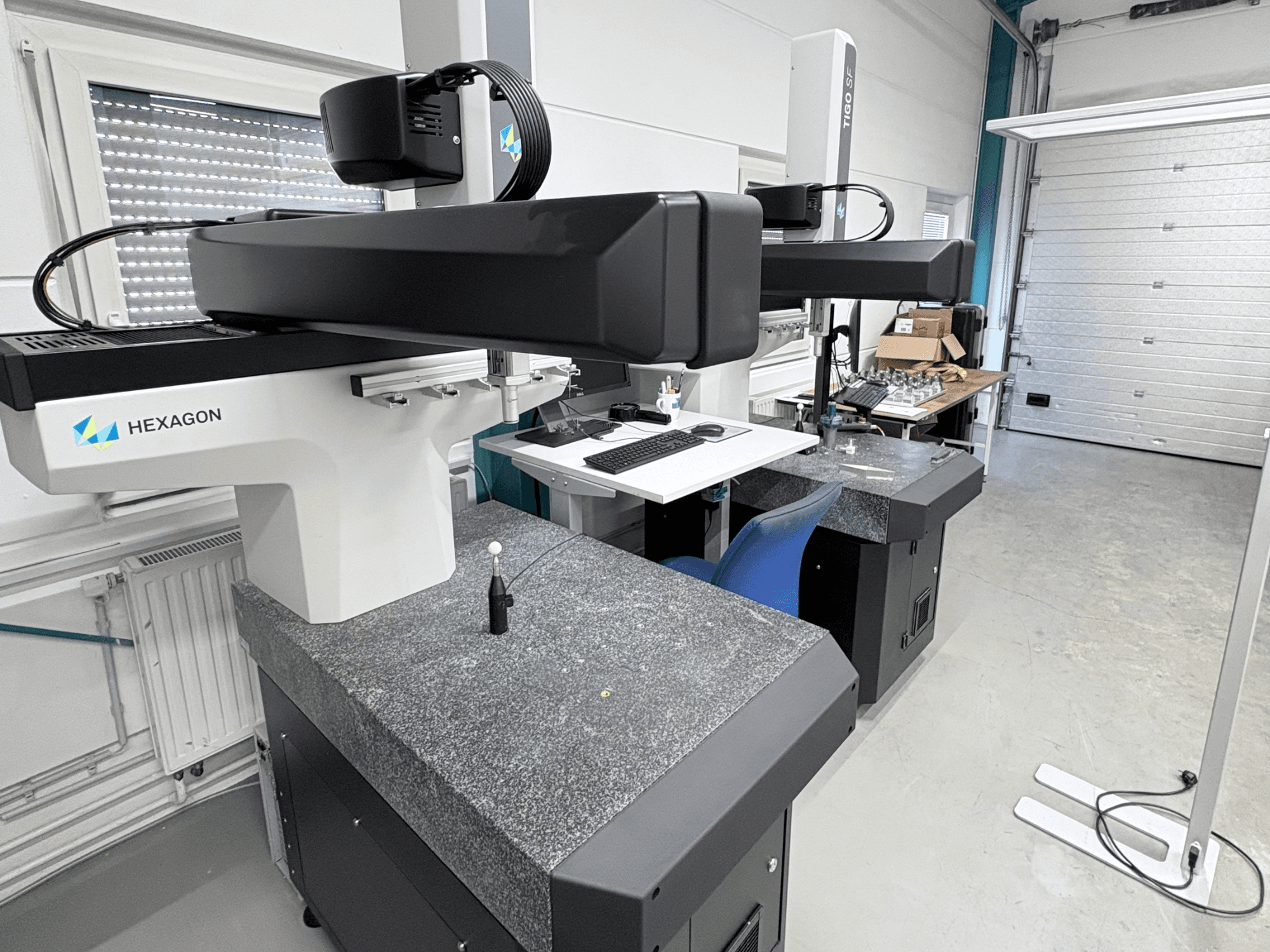Máquina de medição por coordenadas (CMM) Hexagon Tigo SF na oficina, com uma base de granito, sonda de medição e uma estação de trabalho próxima com ferramentas.
