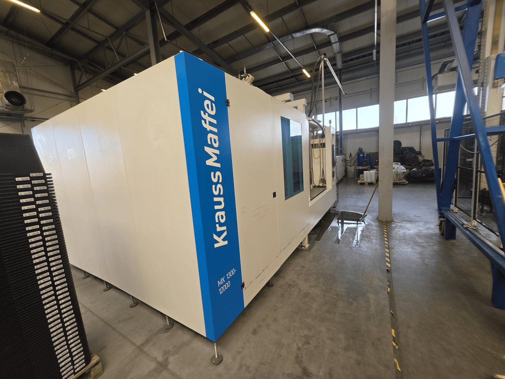 Máquina de moldagem por injeção hidráulica Krauss Maffei MX 1300-12000 vista de lado, mostrando o seu grande chassis branco e a marca azul.
