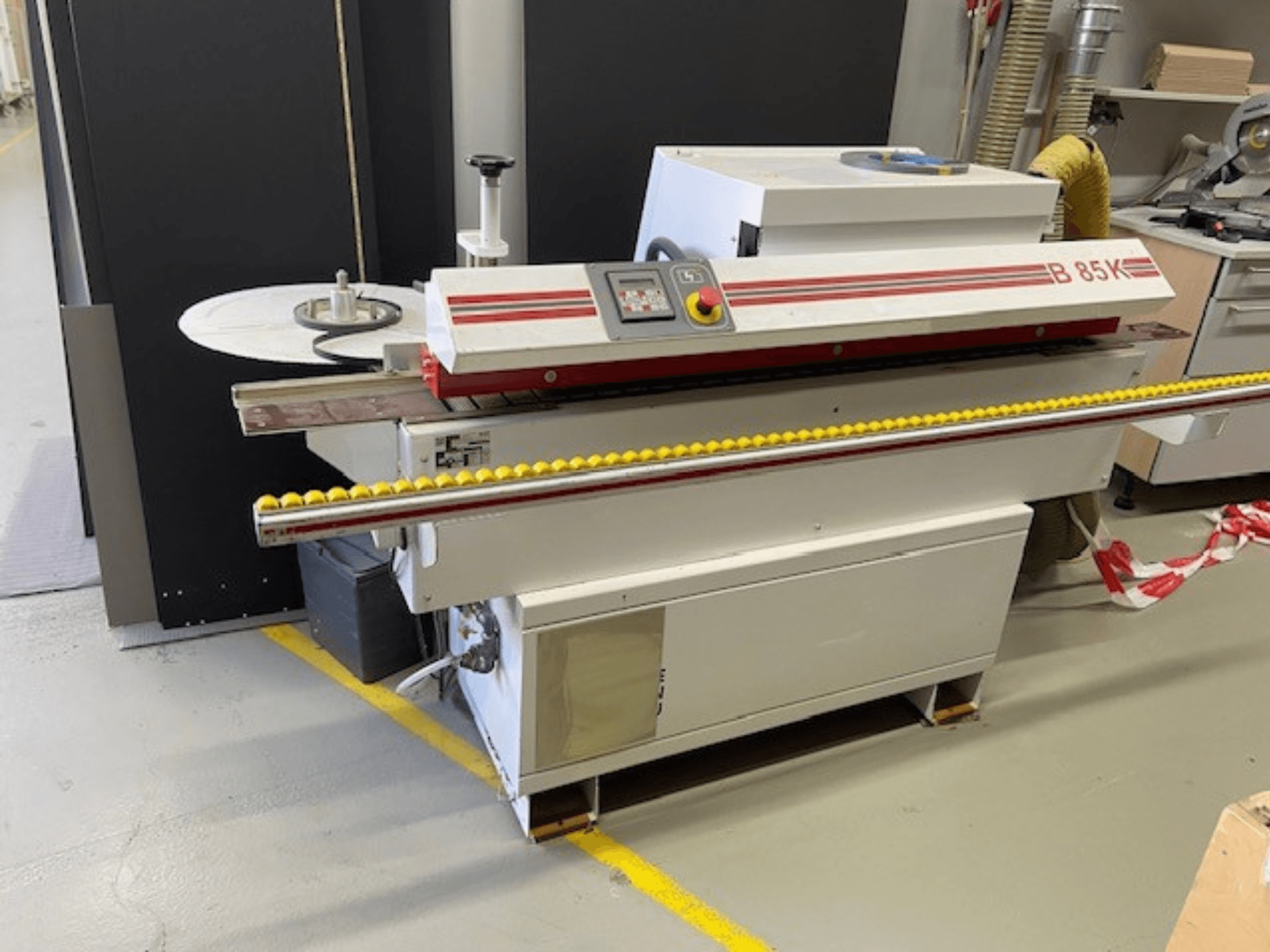A coladeira de bordos B 85K em branco e vermelho, vista frontal, possui um painel de controlo e um rolo amarelo para alimentação do material.