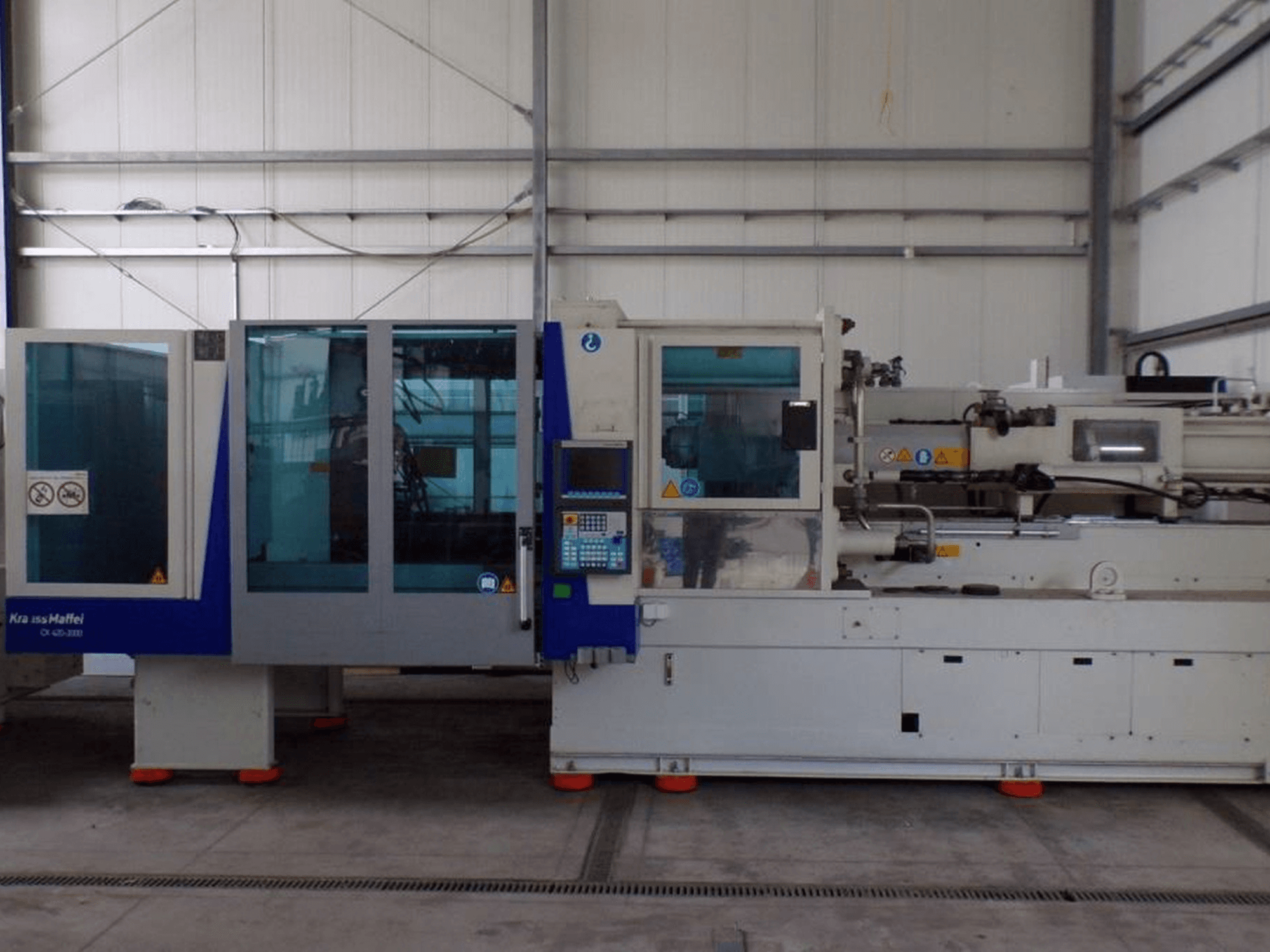 Máquina de moldagem por injeção KraussMaffei, vista lateral, com painel de controlo e indicadores de segurança, visível em fábrica.