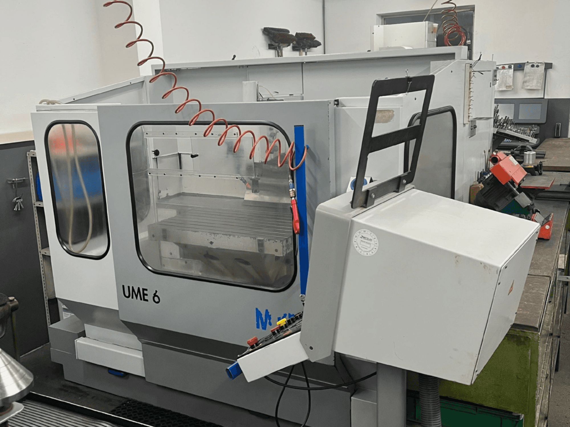 Centro de maquinação universal MIKRON UME600 numa oficina, vista lateral mostrando o painel de controlo e a caixa de proteção.