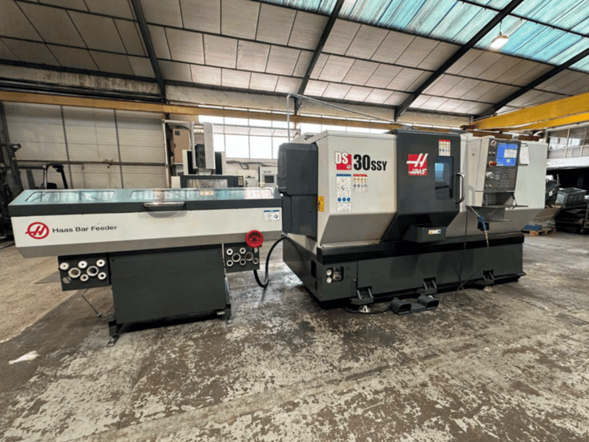 Torno CNC Haas DS-30SSY com alimentador de barras Haas, vista lateral mostrando o painel de controlo e o mecanismo de alimentação num ambiente de oficina.