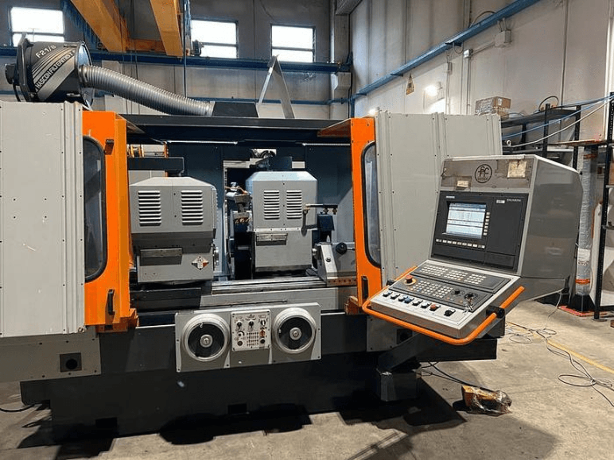 Vista frontal de uma máquina CNC com fusos duplos, painel de controlo e detalhes em laranja. Inclui conduta de ventilação e controlos operacionais.