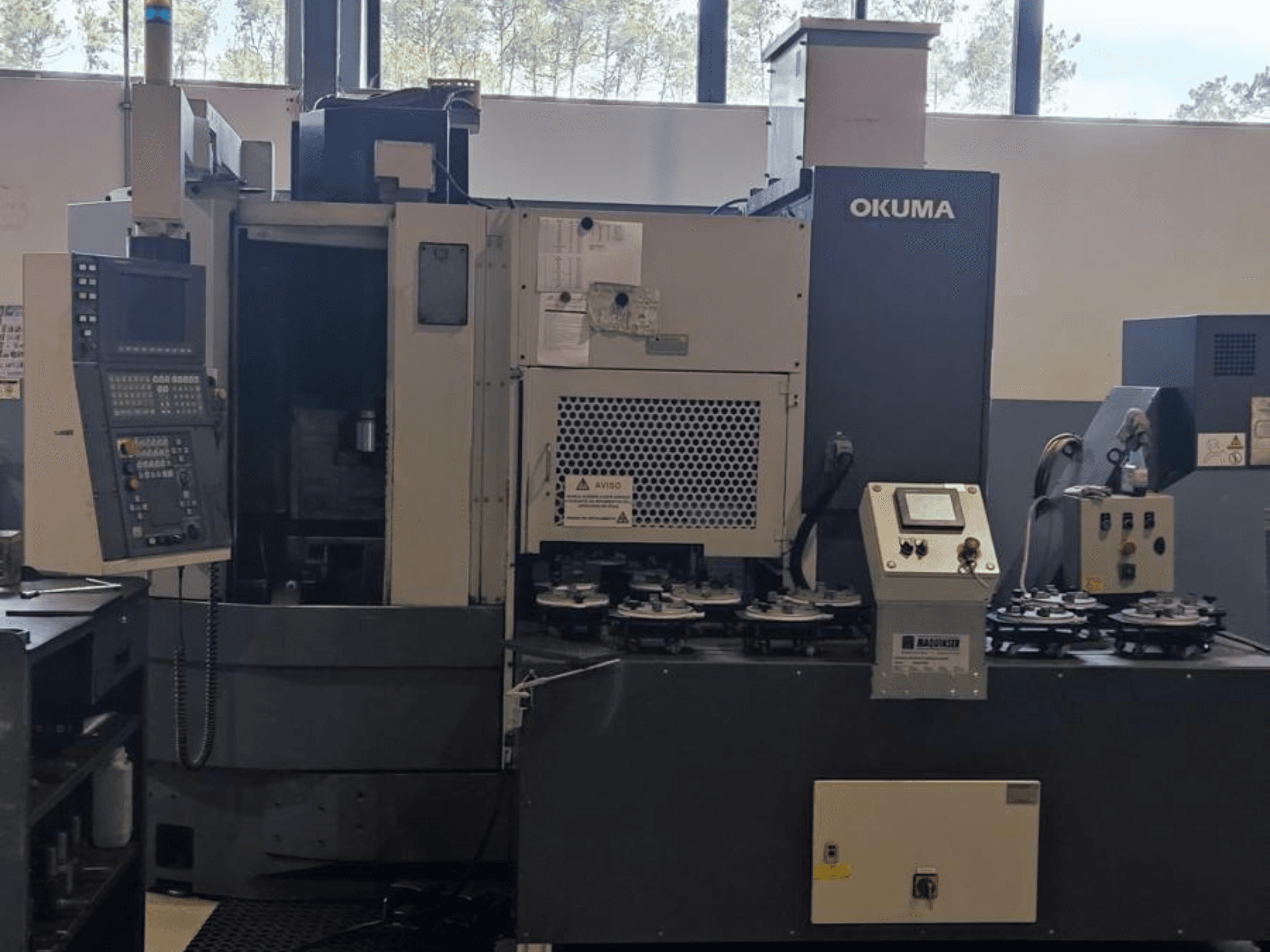 Vista frontal do torno vertical Okuma LVT300, com painel de controlo, suportes de ferramentas e área de maquinação.