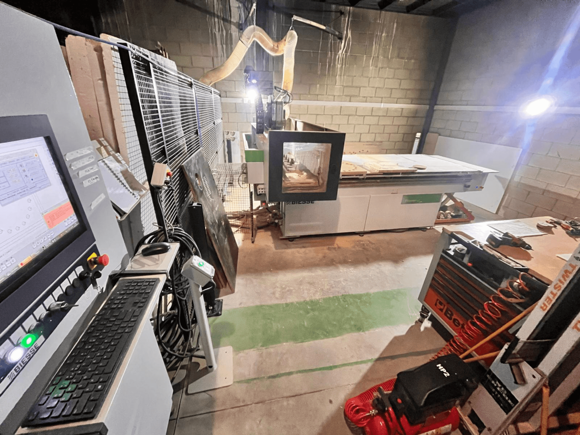 Máquina CNC Biesse numa oficina, vista lateral mostrando o painel de controlo, extração de poeiras, ferramentas e vários materiais sobre a mesa.