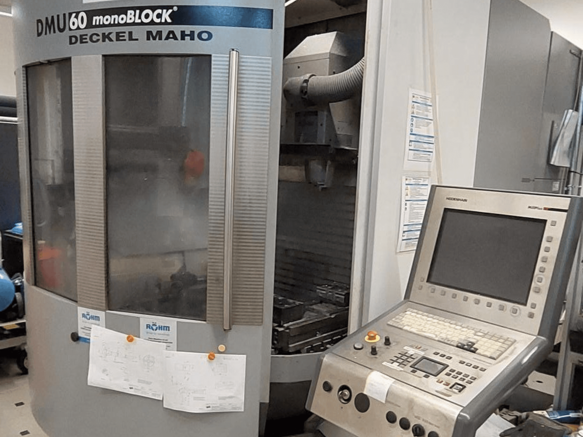 DMU 60 monoBLOCK de Deckel Maho, vista frontal de uma fresadora CNC com um painel de controlo e desenhos técnicos anexados.