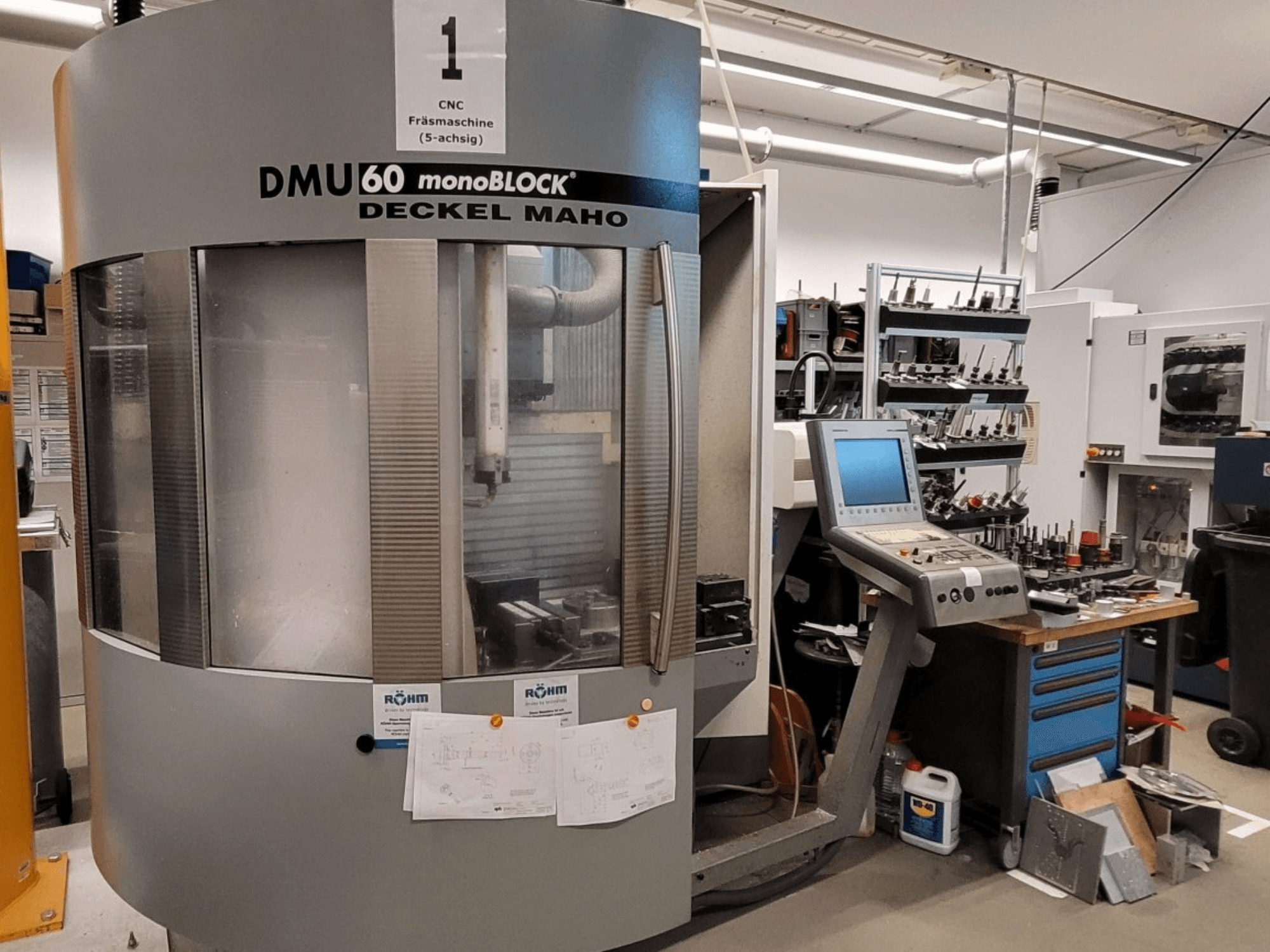 Fresadora CNC DECKEL MAHO DMU 60 monoBLOCK em vista frontal, mostrando o painel de controlo e o armazenamento de ferramentas ao lado.