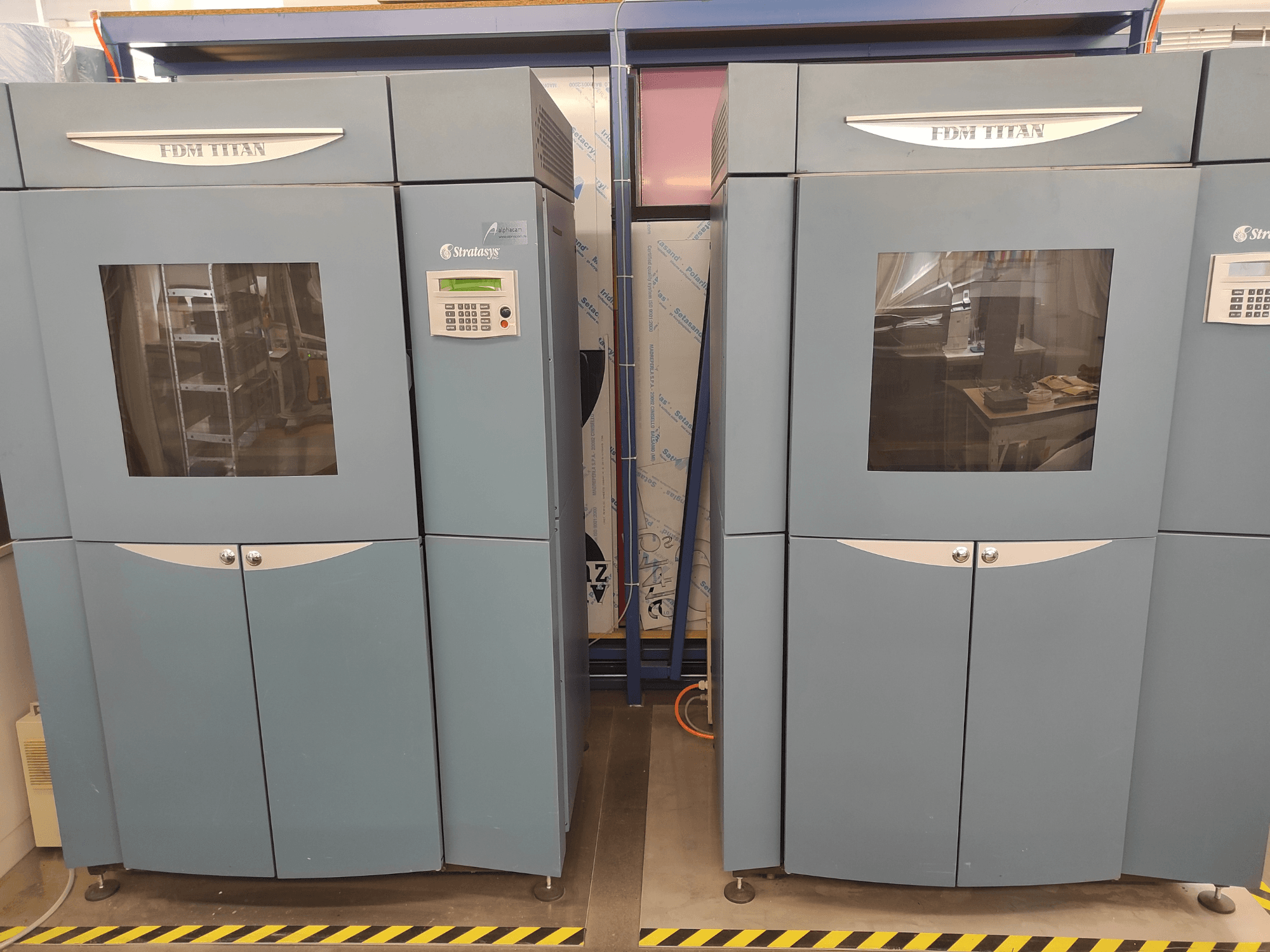 Impressora 3D FDM TitanTI da Stratasys, vista frontal, com portas duplas e painel de controlo, instalada num ambiente industrial.