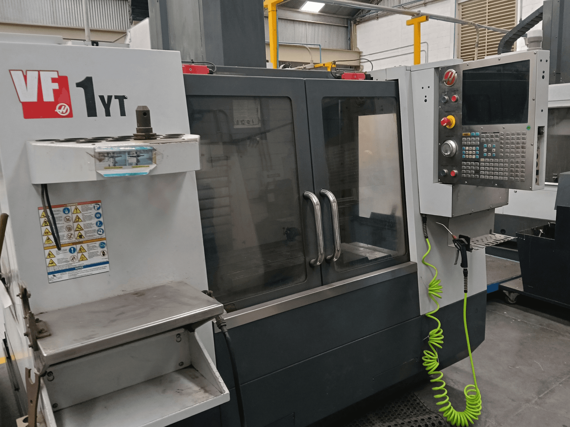 Centro de usinagem vertical HAAS VF-1YT-2, vista frontal, mostrando o painel de controlo, o porta-ferramentas, as etiquetas de segurança e a área de usinagem.