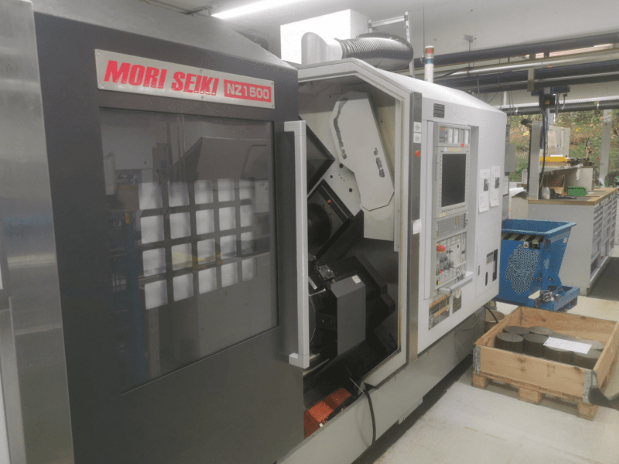 Máquina CNC MORI SEIKI NZ1500 em ligeiro ângulo, mostrando o painel de controlo e o espaço de trabalho com peças metálicas numa palete de madeira.