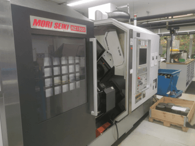 Máquina CNC MORI SEIKI NZ1500 em ligeiro ângulo, mostrando o painel de controlo e o espaço de trabalho com peças metálicas numa palete de madeira.