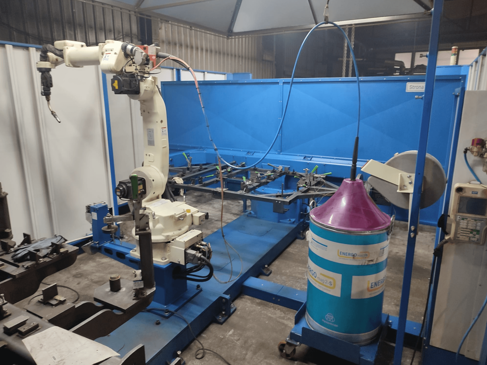 Braço robótico industrial com uma base branca e azul, montado numa oficina com uma superfície de trabalho azul e componentes metálicos.