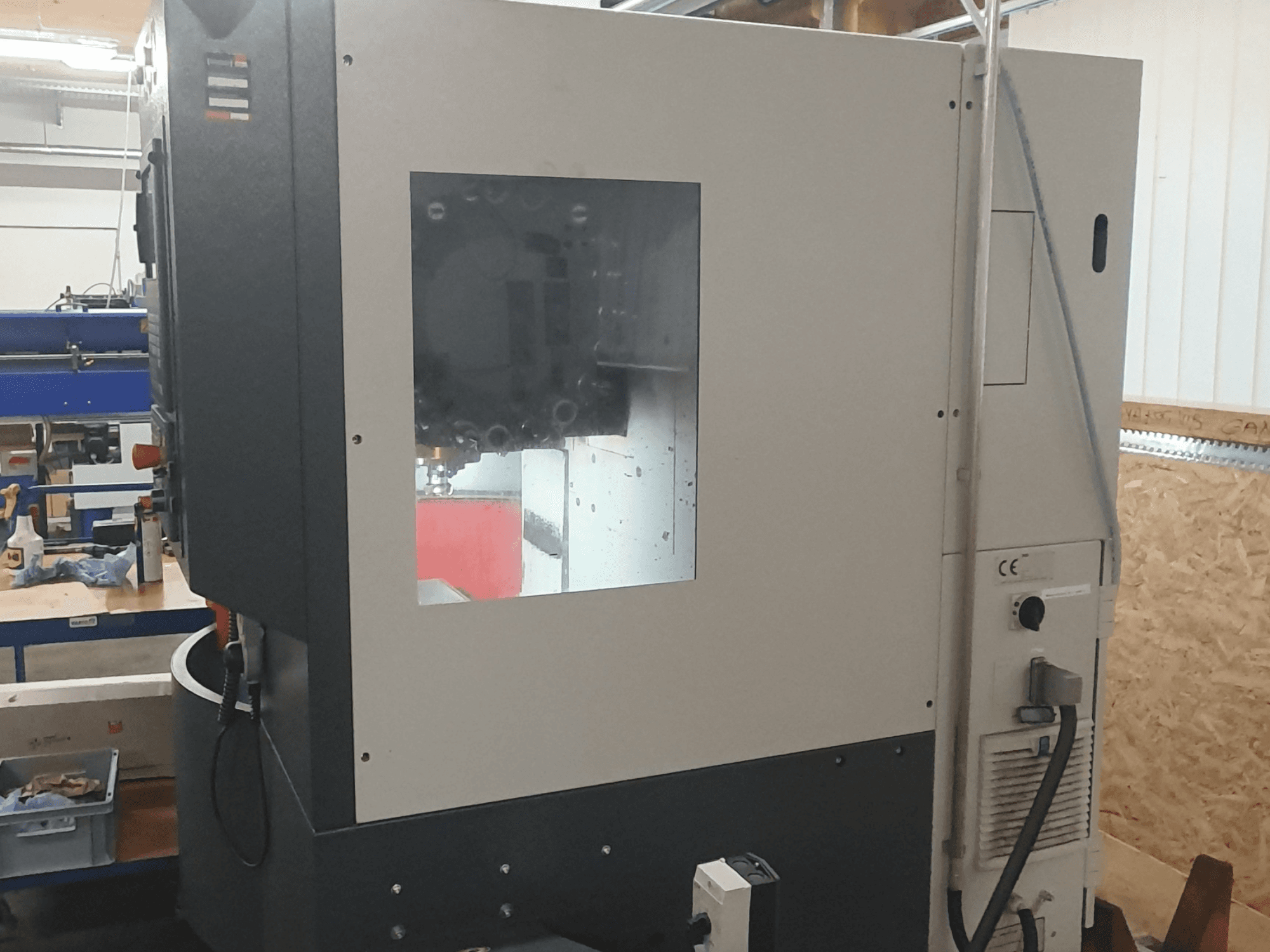 Vista lateral de uma máquina para trabalhar metais com painel de controlo visível, grande janela de visualização e componentes internos complexos.