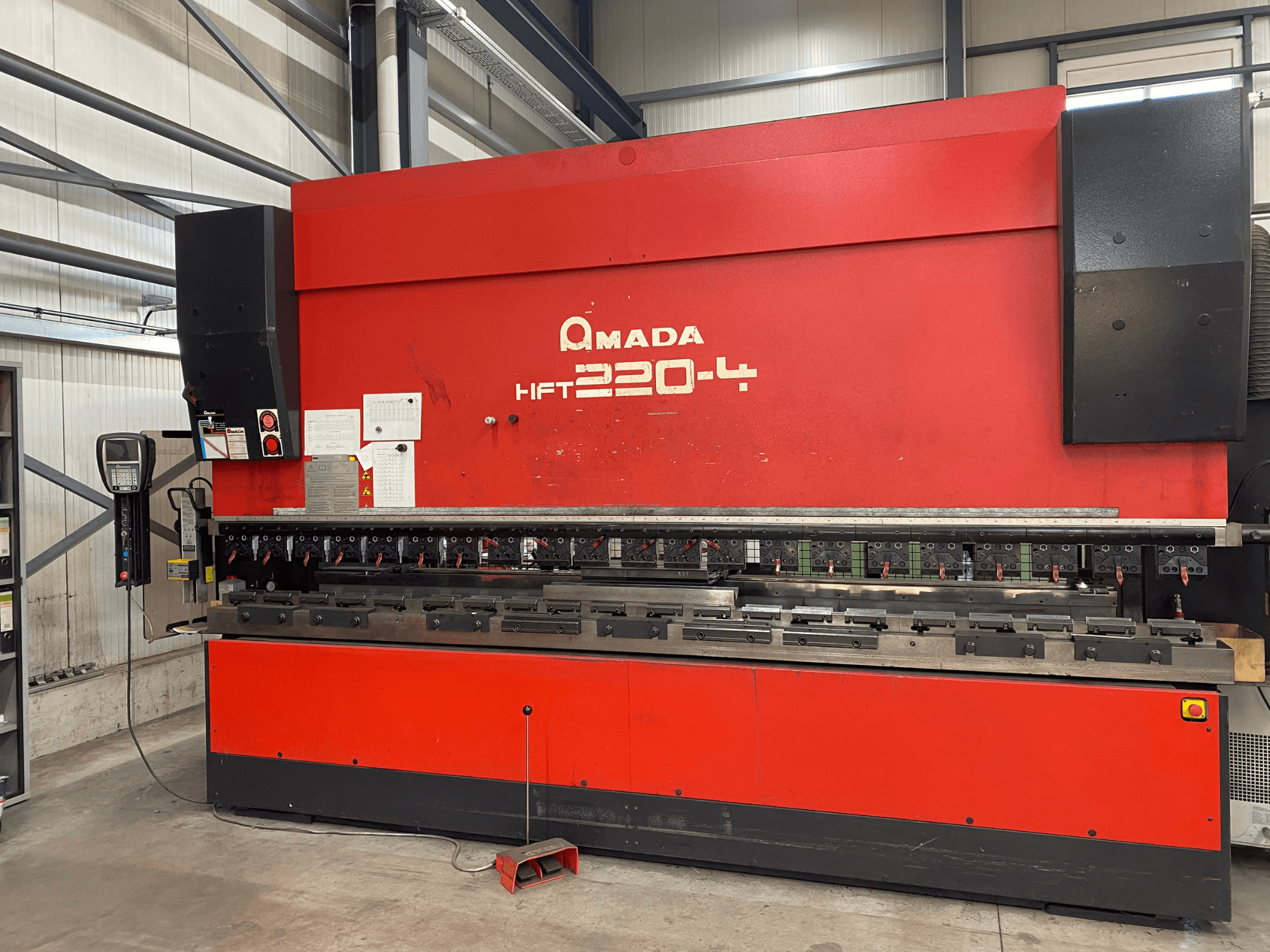 Prensa dobradeira AMADA HFT 220-4 vermelha numa configuração de fábrica, vista frontal mostrando o painel de controlo e as ferramentas de dobragem na superfície de trabalho.