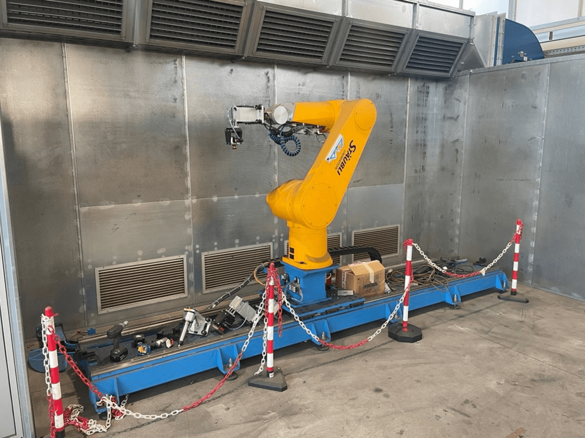 Braço robótico STAUBLI CS8C-RX160 numa oficina, vista lateral, com cabos e várias ferramentas numa base azul rodeada por barreiras de proteção.