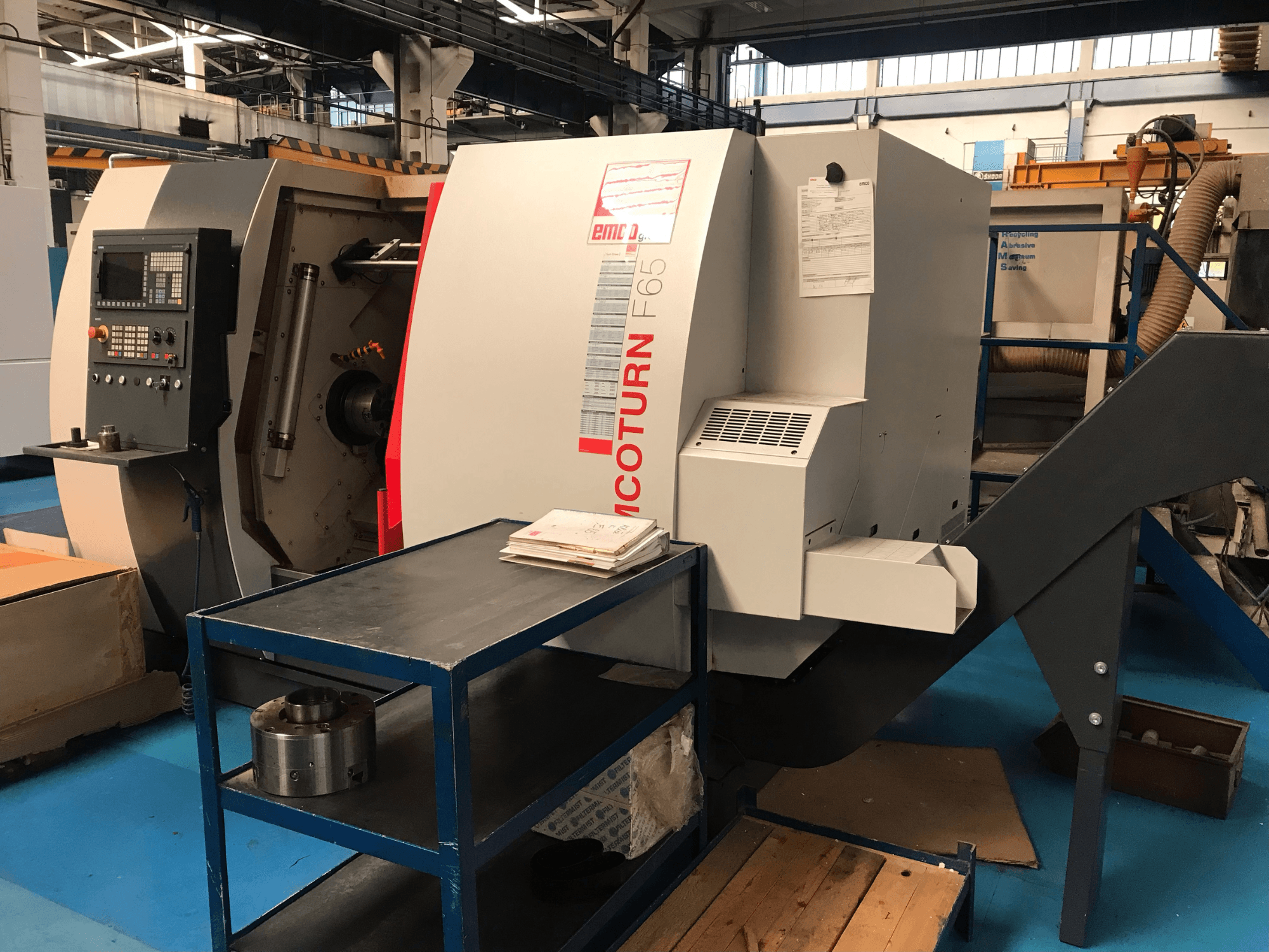 Torno horizontal EMCO Emcoturn 65, visto de lado, mostrando os comandos e a área de trabalho, com peças metálicas nas proximidades.