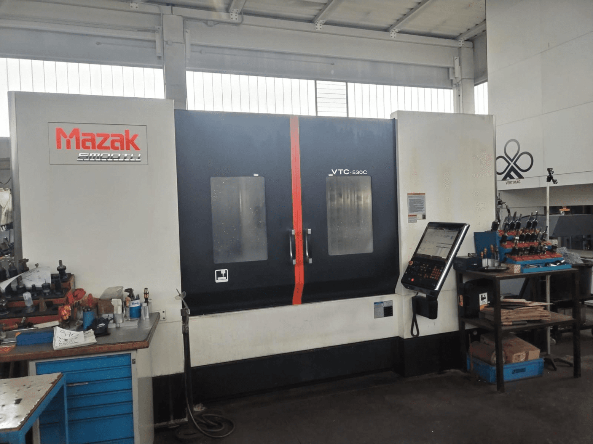 Centro de maquinagem vertical Mazak VTC-530C numa oficina, com um painel de controlo e ferramentas numa mesa lateral.