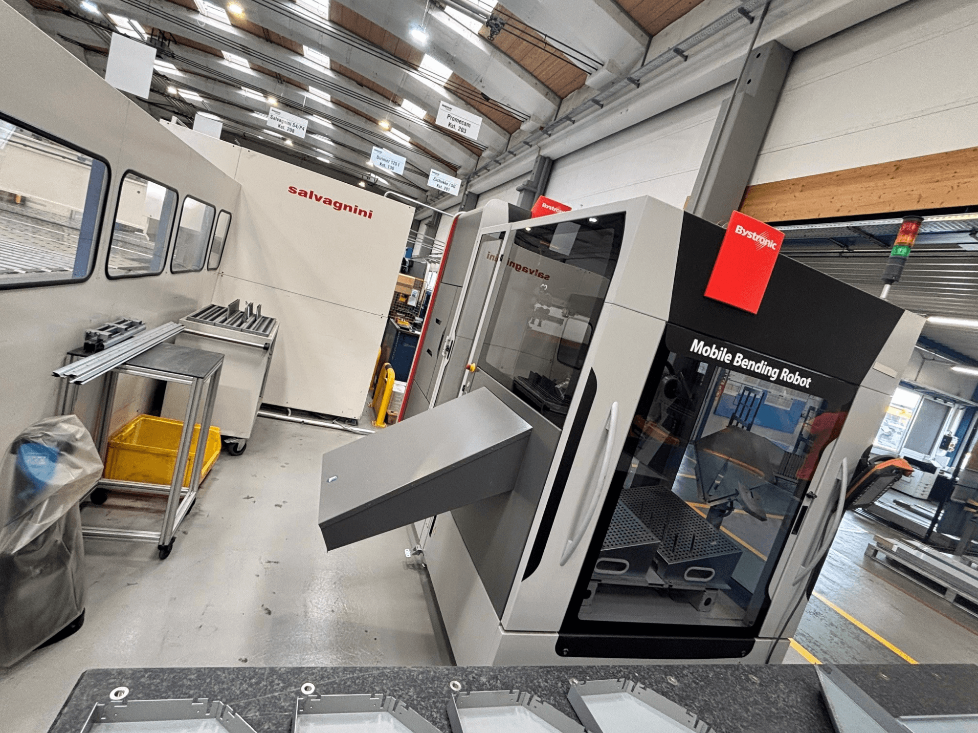 Prensa-dobra Bystronic Xpert 40/1030, vista frontal que mostra o robô de dobragem móvel e a área de trabalho com peças metálicas sobre uma mesa.