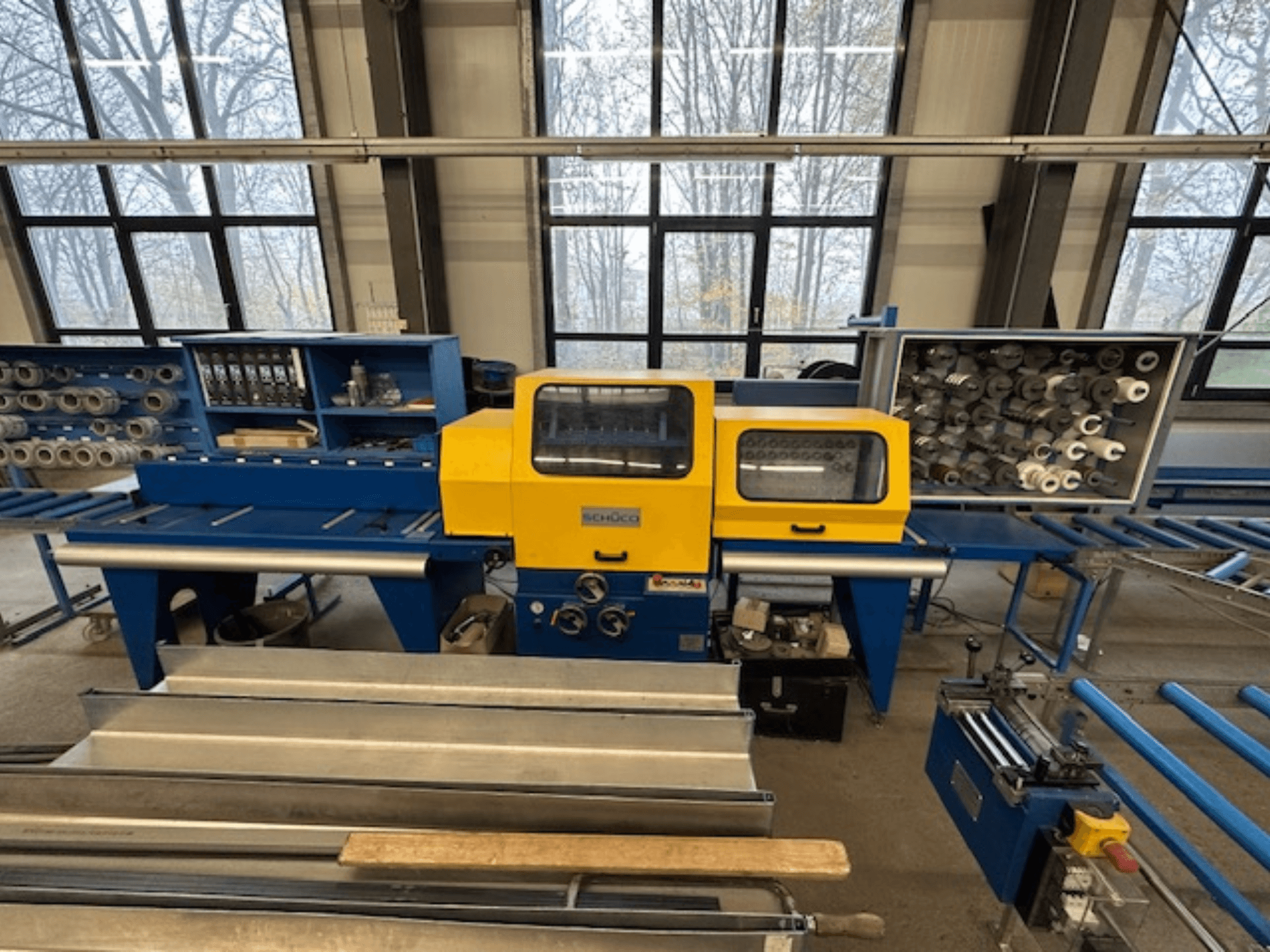 Vista frontal da máquina-ferramenta SCHÜCO ISOMAT 4, com um painel de controlo amarelo, vários rolos e um compartimento de arrumação.