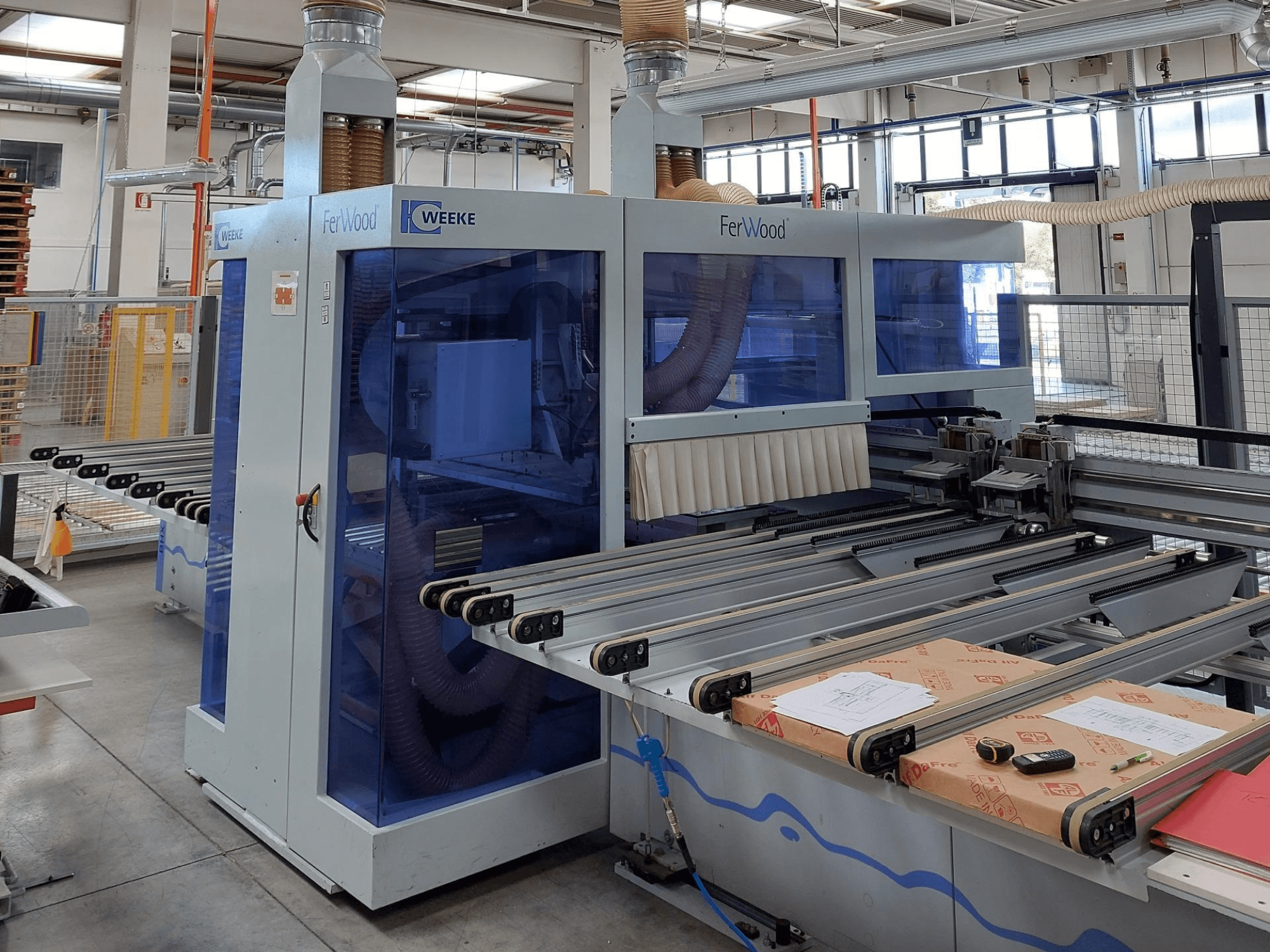 Máquina CNC Weeke FerWood numa oficina, vista frontal mostrando painéis azuis, botões de controlo e mesa de trabalho com materiais.