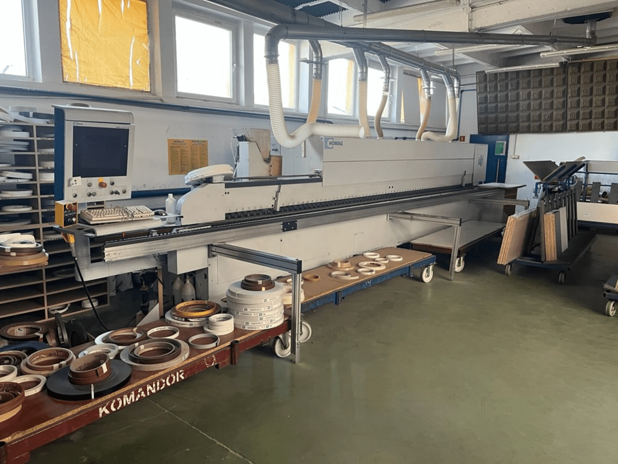 Máquina de aplicação de orlas HOMAG numa oficina, com comandos e vários materiais de aplicação de orlas num carrinho azul. Vista lateral.