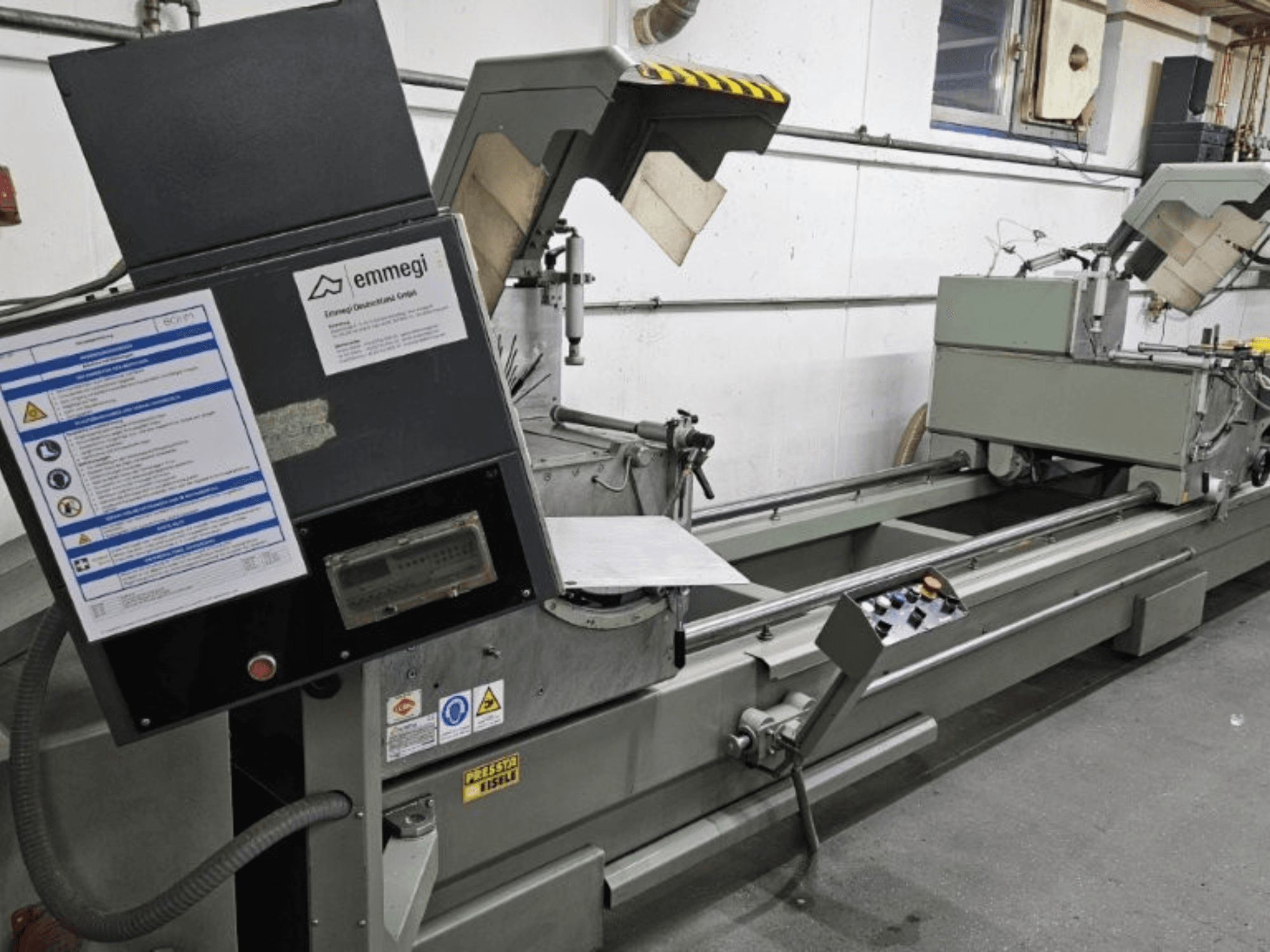 Máquina de corte industrial Emmegi, vista frontal, com painel de controlo, etiquetas de segurança e componentes operacionais.