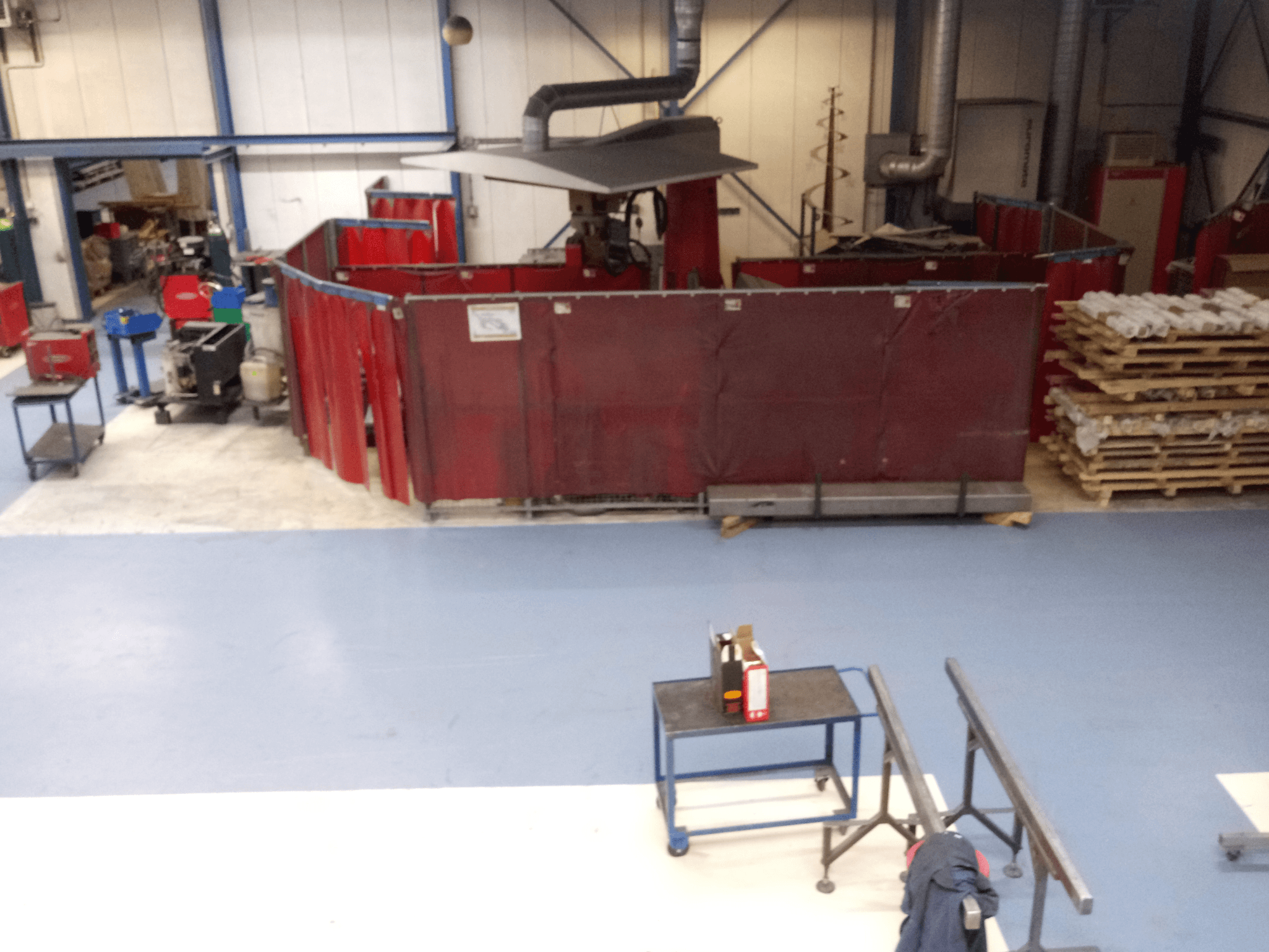 Espaço de trabalho industrial com cortinas de soldadura vermelhas, máquinas e paletes; vista aérea mostrando a disposição e o equipamento.