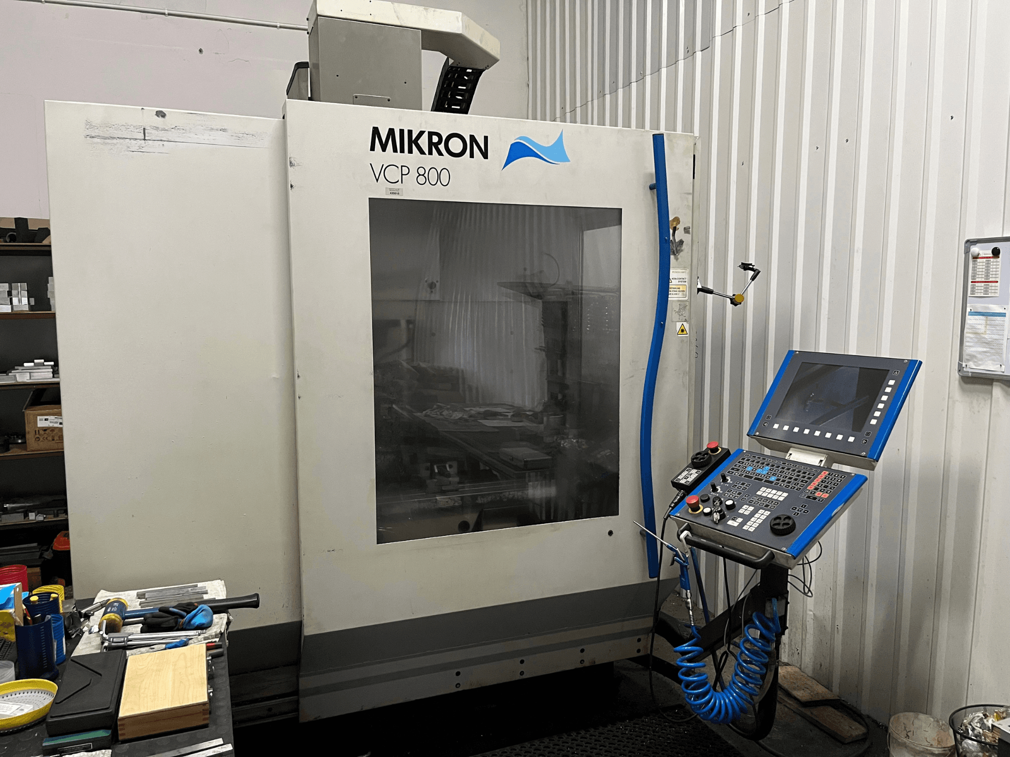 Centro de maquinagem vertical MIKRON VCP 800, vista frontal, com o painel de controlo e as ferramentas numa bancada de trabalho em primeiro plano.
