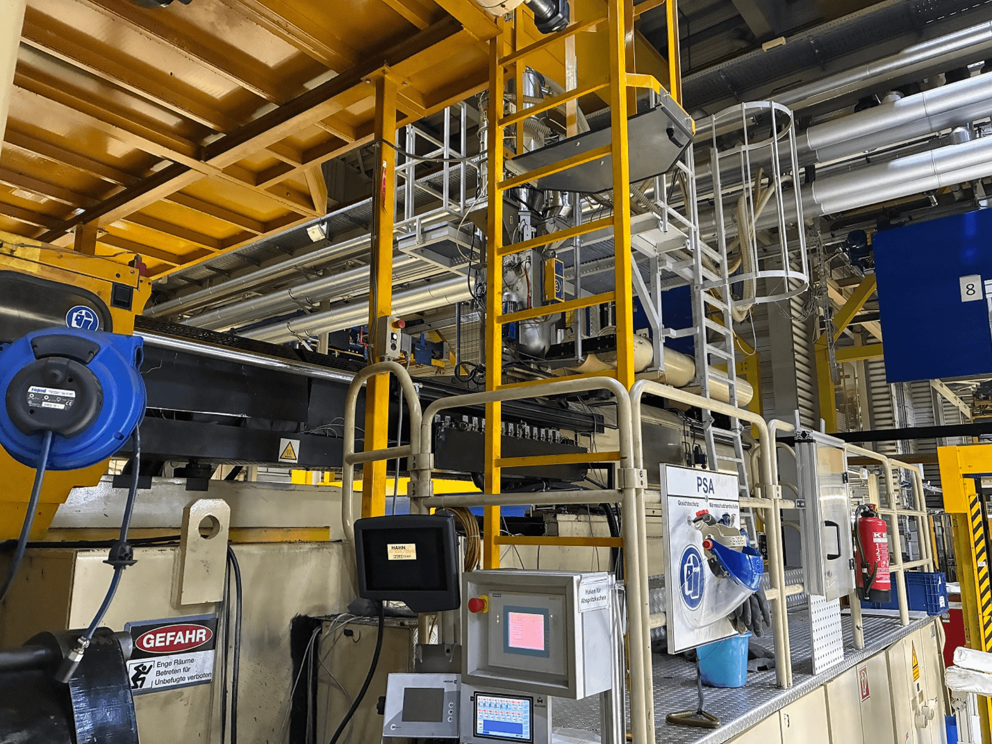 Painel de controlo de máquinas industriais com equipamento de segurança azul e andaimes amarelos, vista lateral mostrando tubos e sinais de aviso.